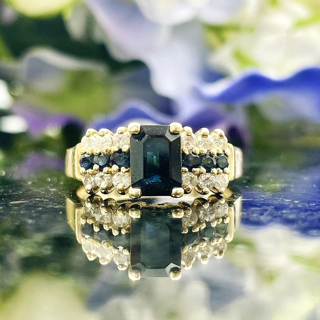 close up in front of flowers Emerald Cut Sapphire With Diamond And Sapphire Accents Ladies Ring 14k Yellow Gold