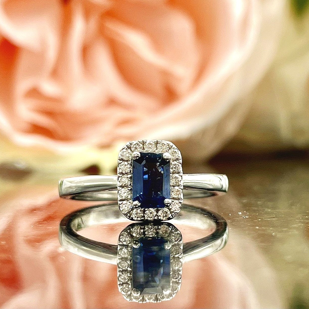 close up in front of flowers .64ct Emerald Cut Sapphire With Diamond Halo Ladies Ring 14k White Gold