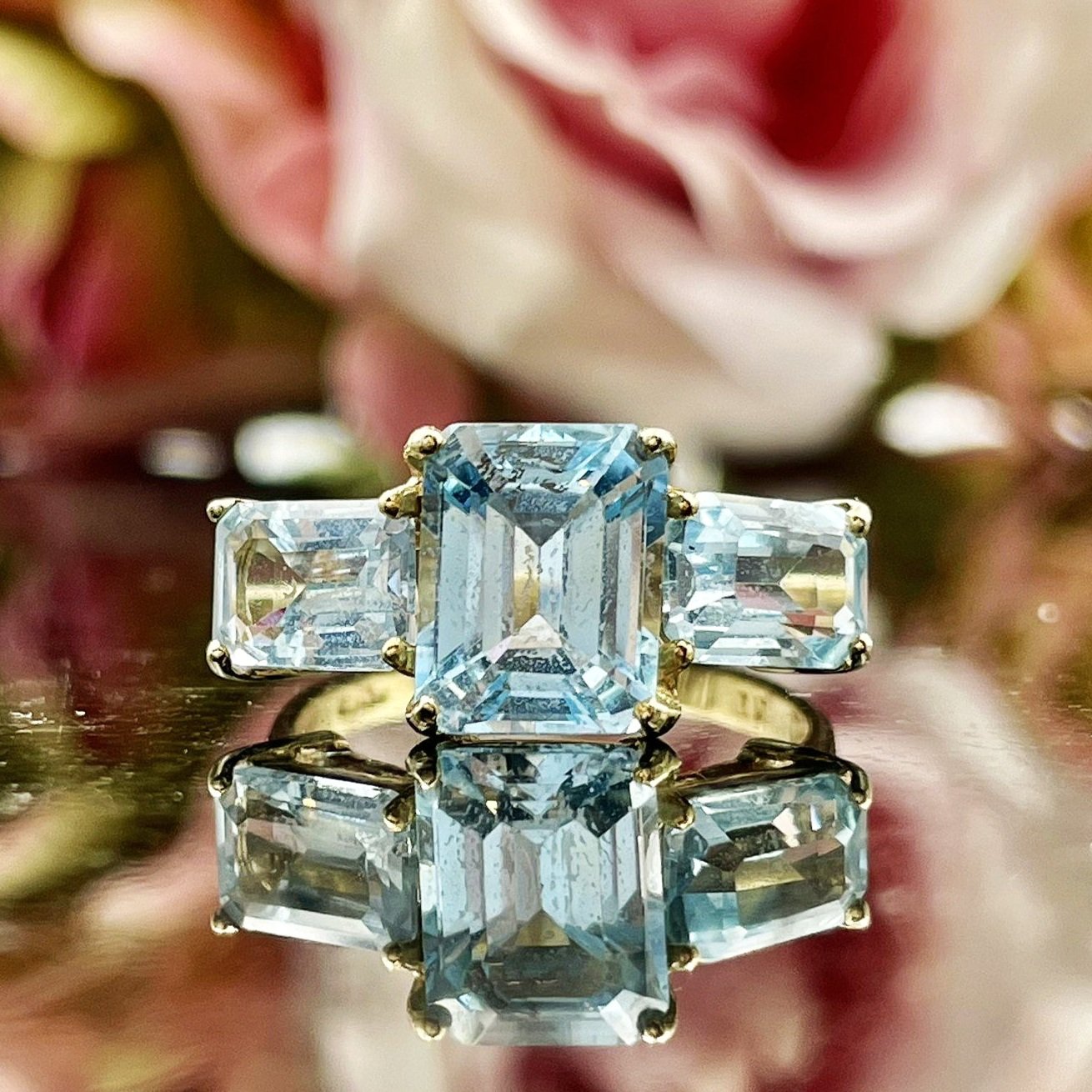close up in front of flowers 10k Yellow Gold Emerald Cut Blue Topaz Three Stone Ring