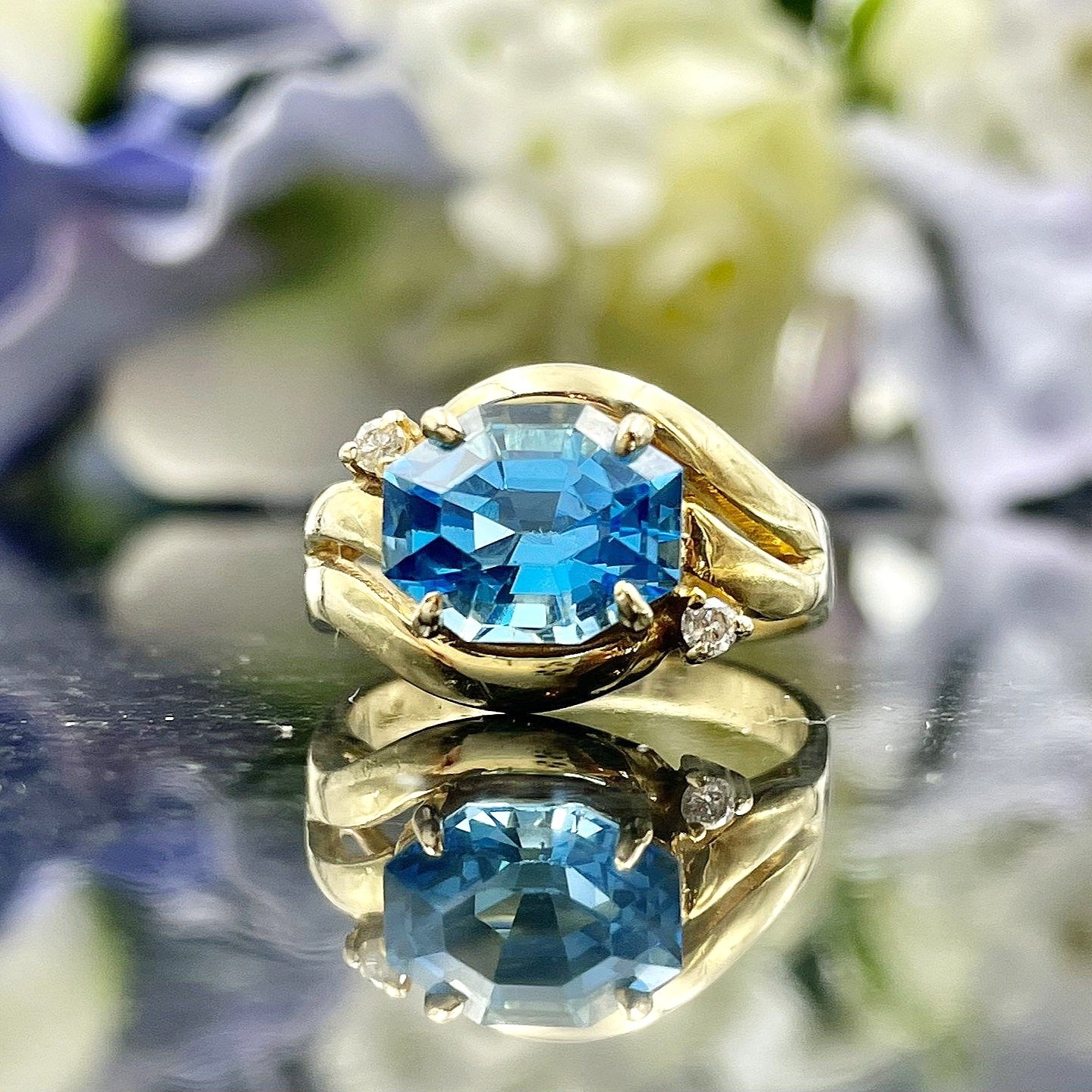 close up in front of flowers Octagonal Cut Blue Topaz With Diamond Accents Swirl 14k Yellow Gold Ladies Ring