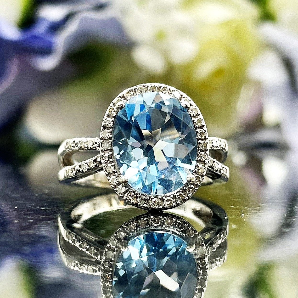 close up in front of flowers Oval Blue Topaz With Diamond Halo And Split Shoulders 14k White Gold Ring