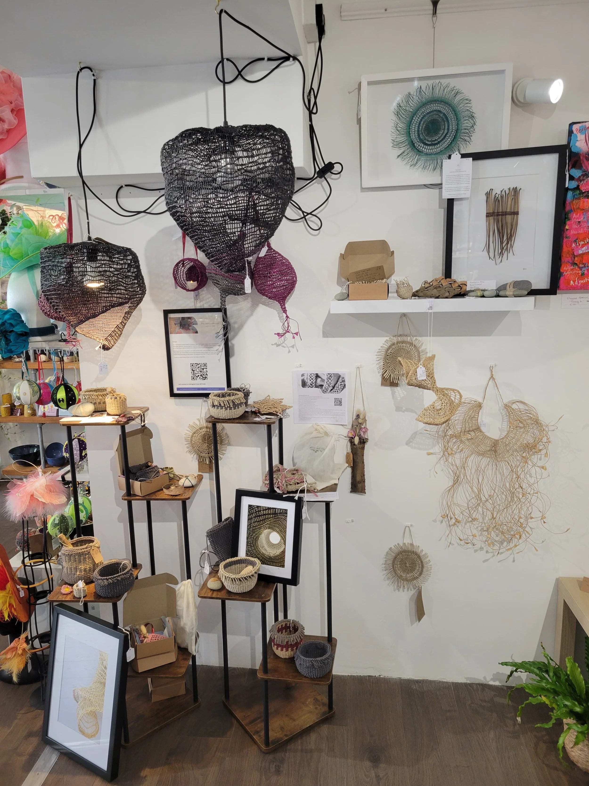 Display of various woven art pieces, including baskets, wall hangings, and sculptures, arranged on shelves, frames, and walls in a craft store.