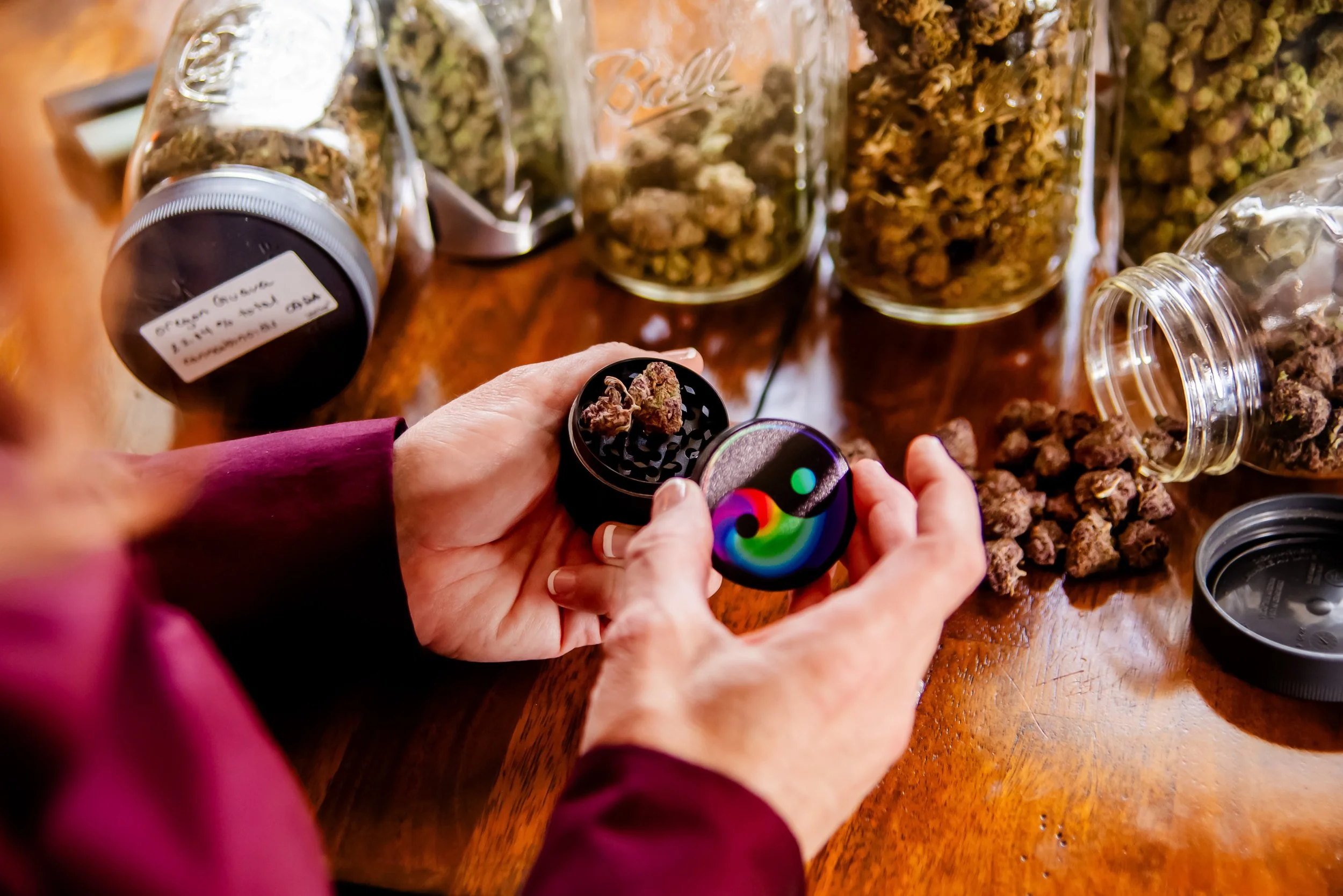 Person holding a grinder with cannabis buds, surrounded by jars and containers filled with flower on a wooden table.