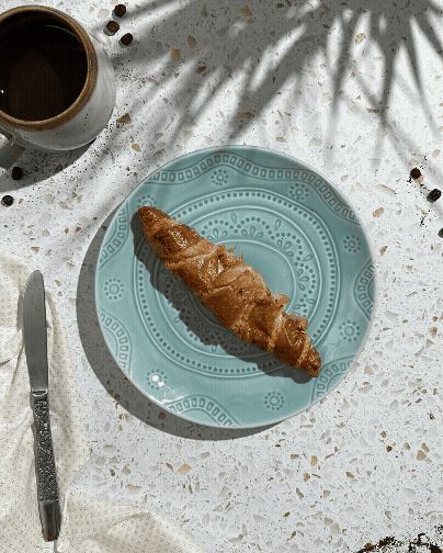 A croissant on a blue patterned plate with a cup of coffee nearby on a white speckled table with coffee beans and shadows of plant leaves.