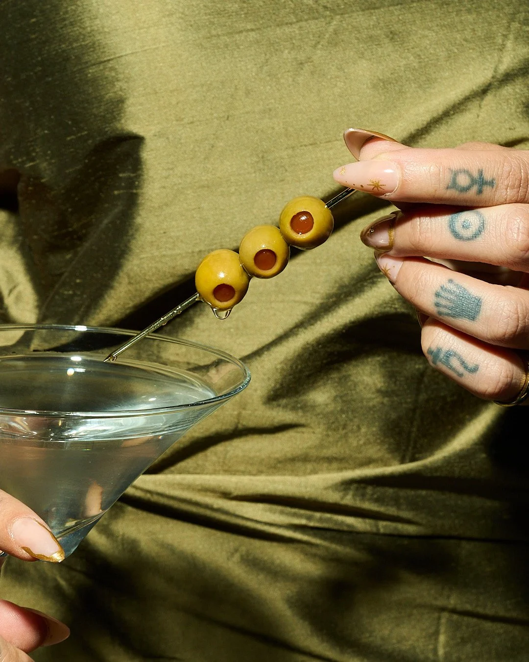 Close-up of a hand with tattoos holding a glass martini and a skewer with green olives over a green fabric background.