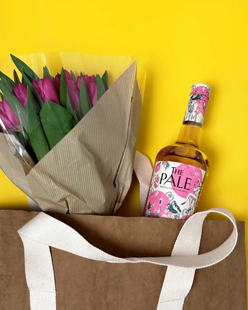 A paper bag containing pink tulips with green leaves, a pink bottle of wine with a label reading "The Pale," and a yellow background.
