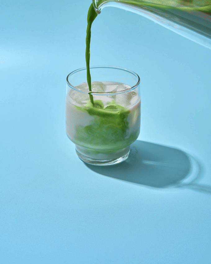 Green juice being poured into a glass with ice and cucumber slices, on a light blue background.