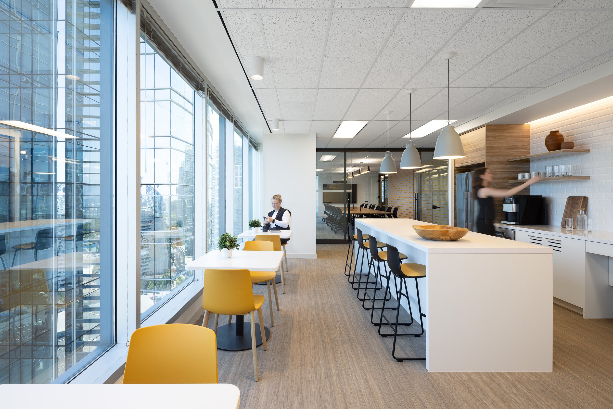 Sun Life office kitchen and seating area in Burnaby with floor-to-ceiling windows, yellow chairs, a long island with bar stools, and staff using the space.