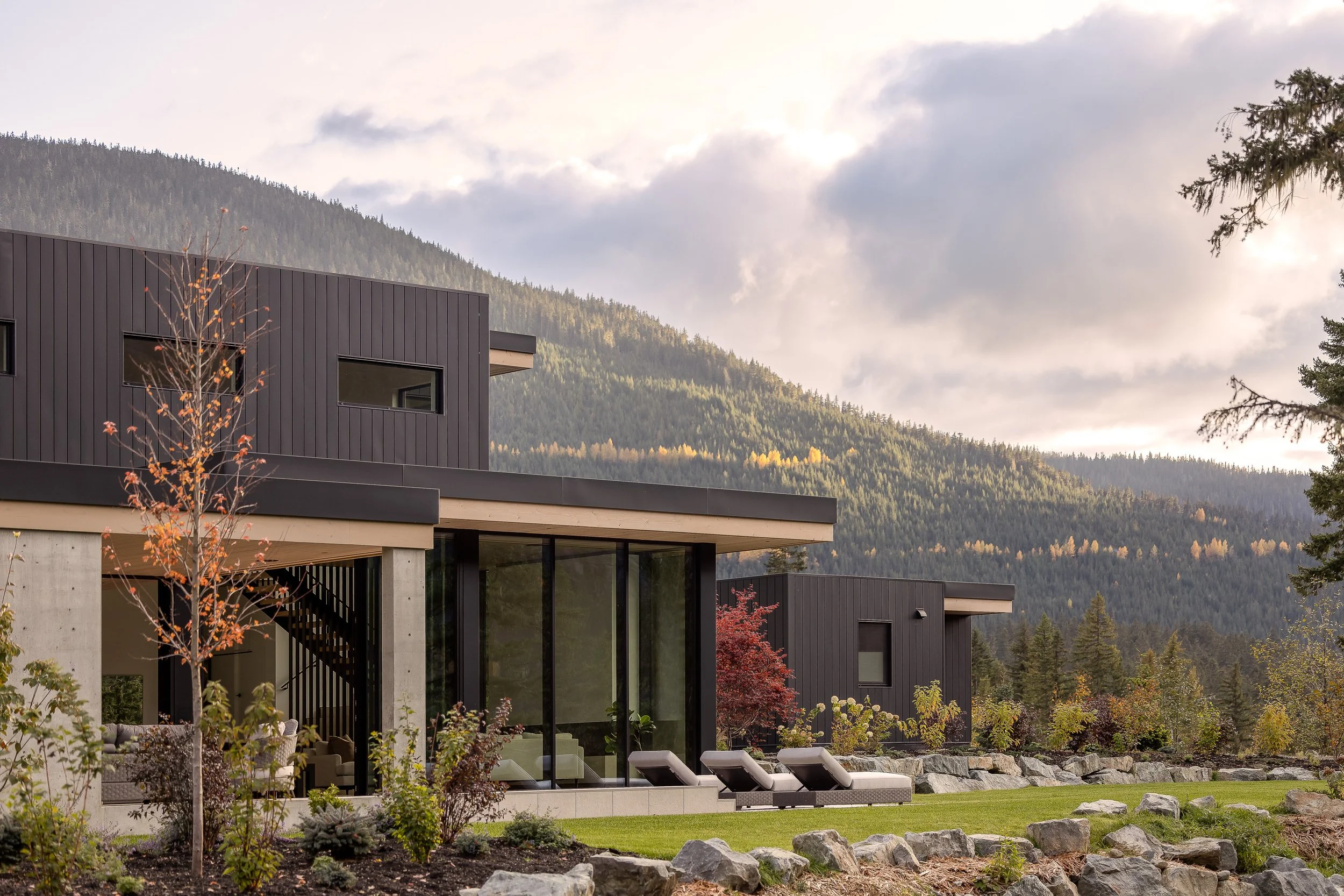 Exterior side/back: glazing, patio loungers, landscaping/rocks, soft sky + mountain.
