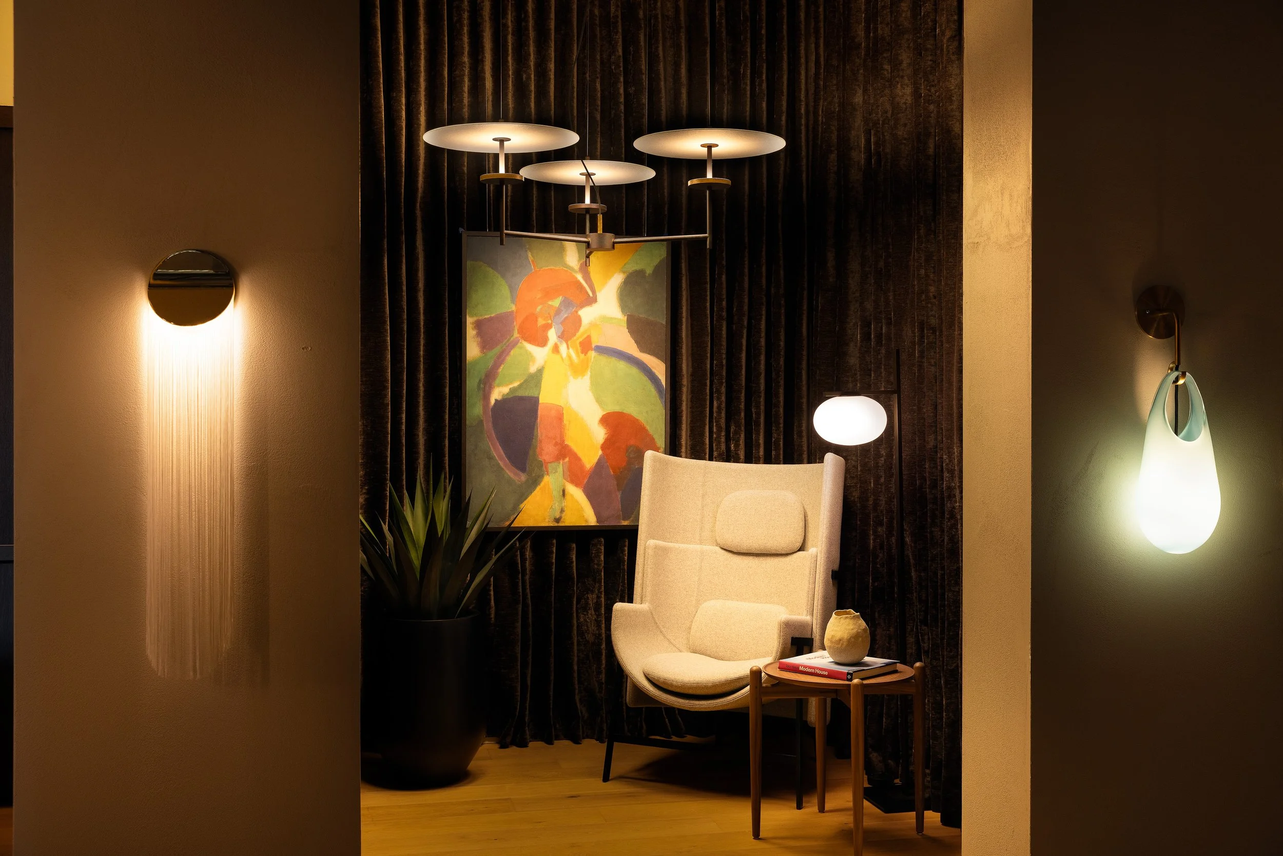 Showroom seating vignette with an accent chair, abstract artwork, glowing wall sconces, and a modern multi-disc chandelier.