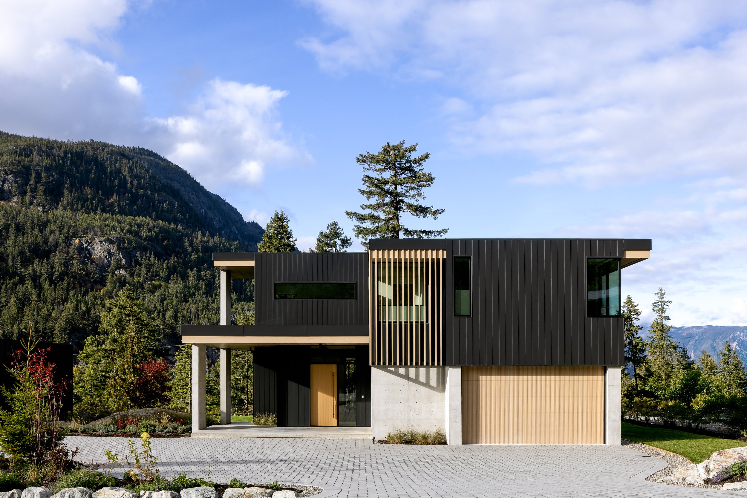  Front elevation / street-side hero: black cladding + vertical wood screen, mountain backdrop, blue sky.