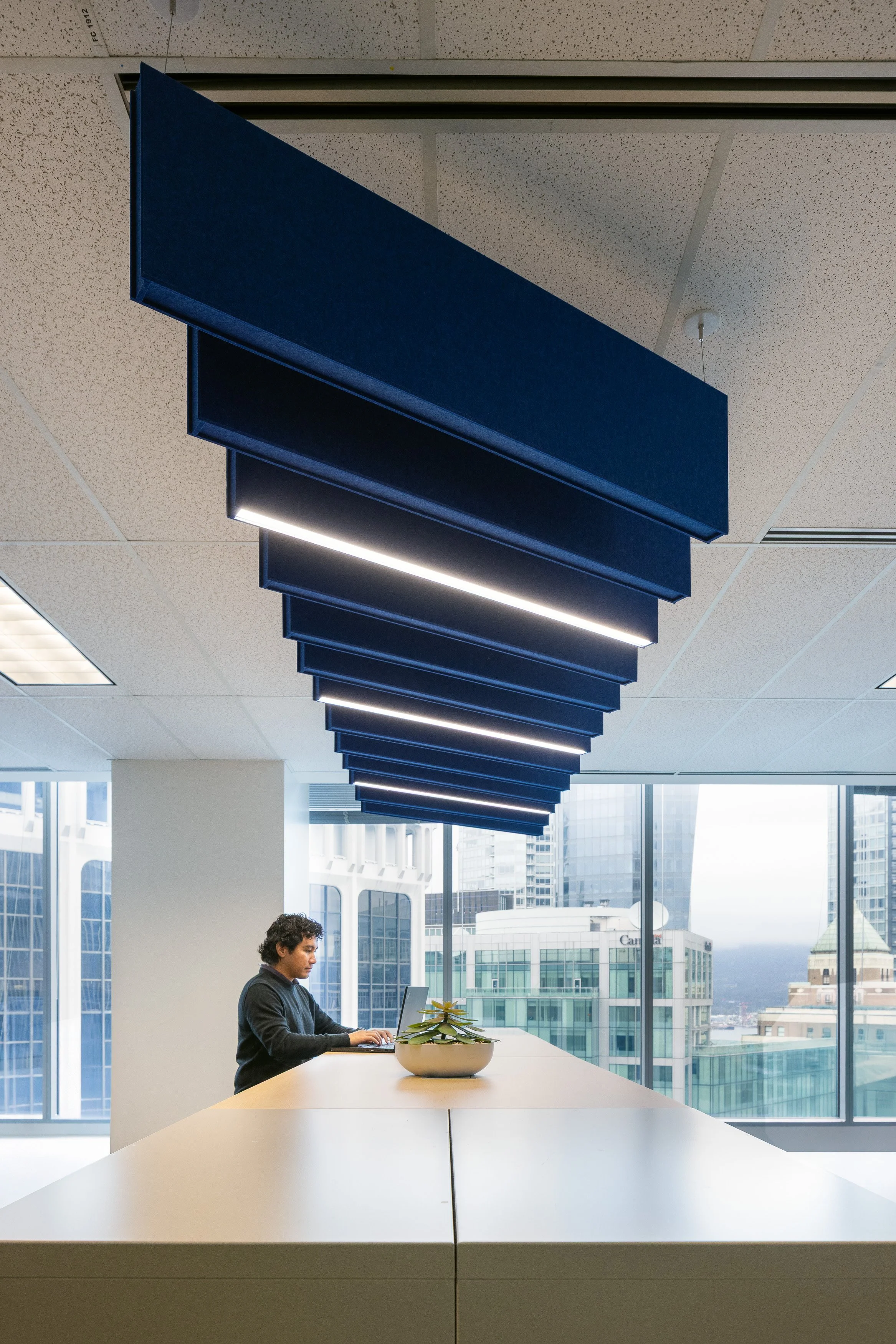 Employee working at a shared high table beneath custom acoustic ceiling baffles at the Mott MacDonald Vancouver office.