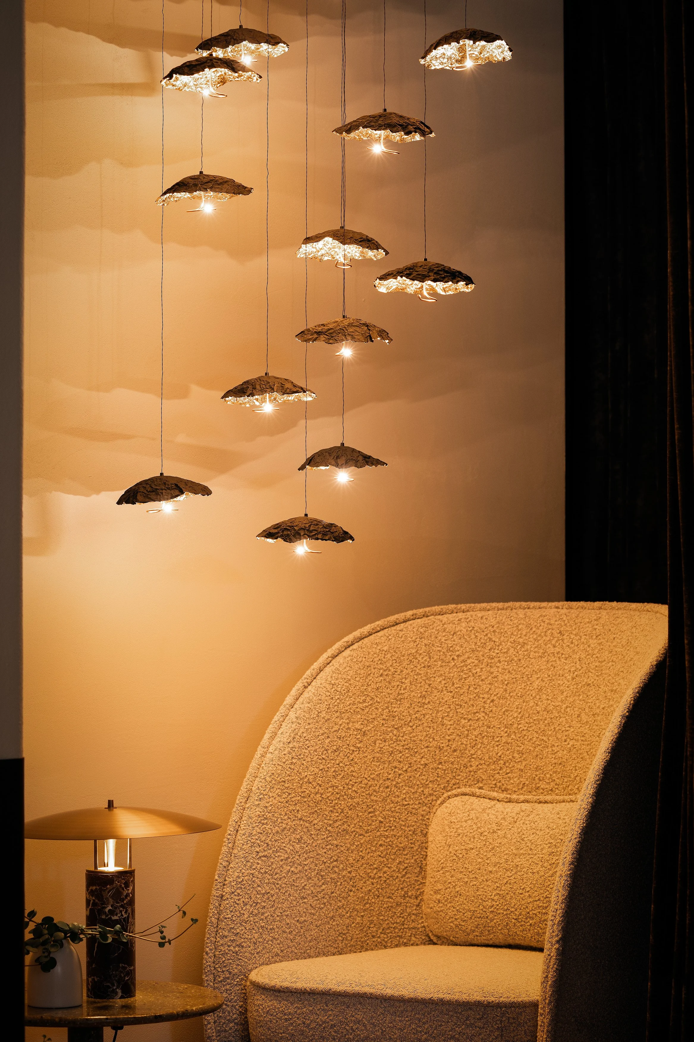 Textured lounge chair beside a small side table and lamp, with a cluster of sculptural hanging pendants glowing overhead.