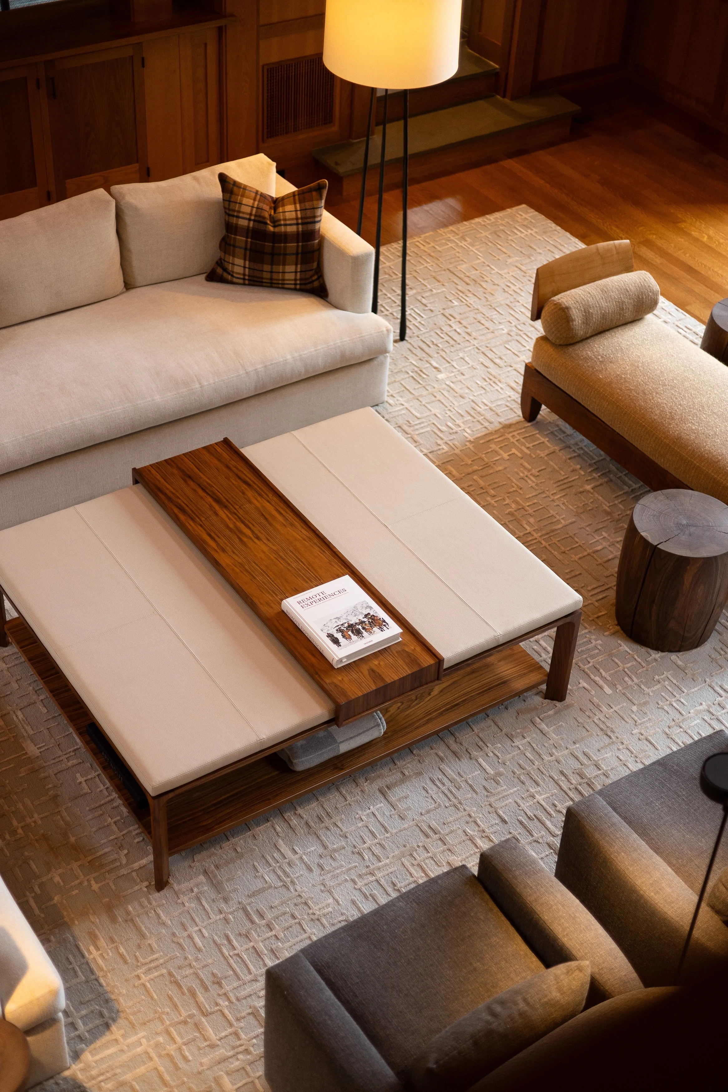A cozy living room with a white sofa, plaid pillow, beige lounge chair, wooden side table, floor lamp, and a coffee table with a stack of magazines on top, all on a textured beige rug.
