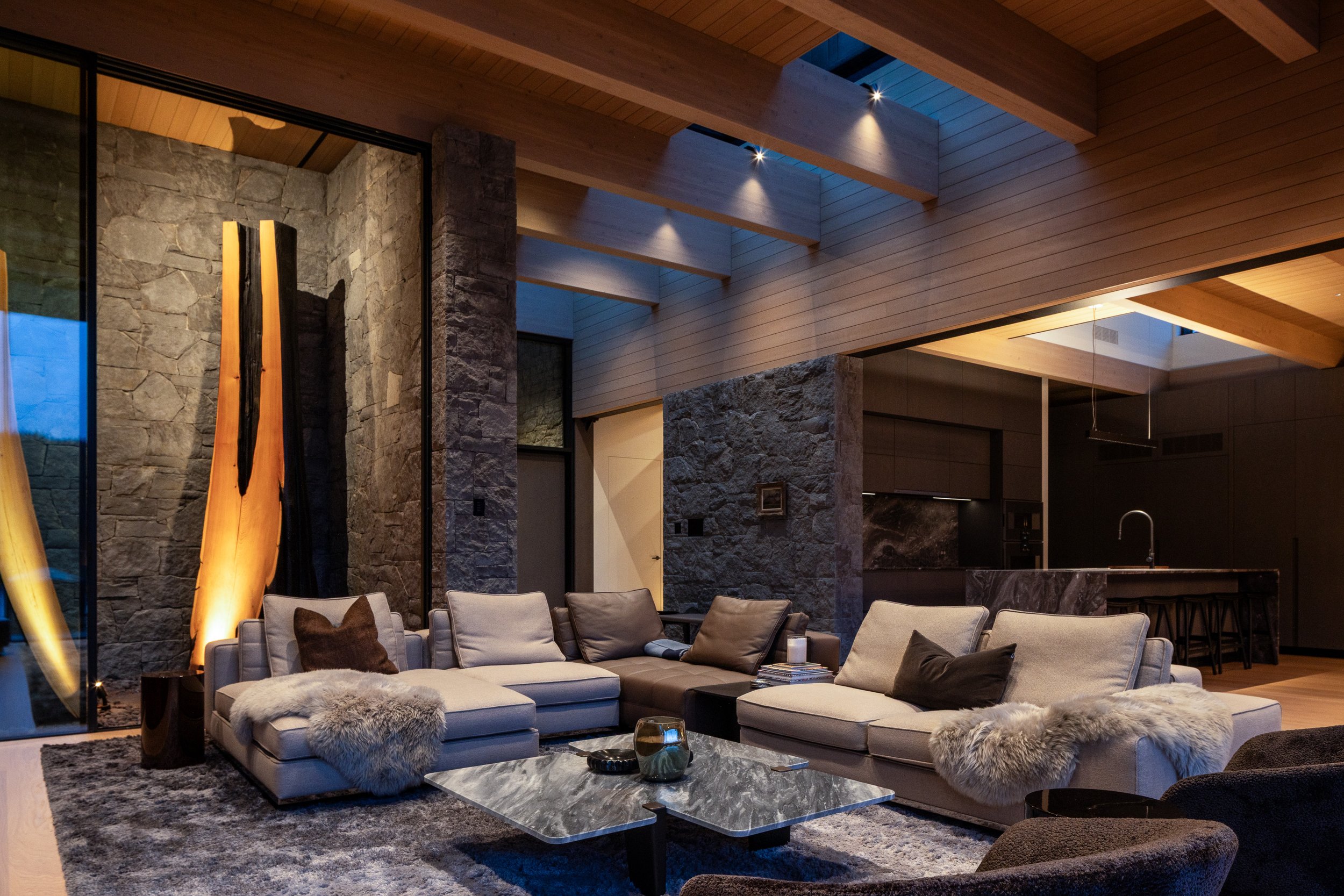 An evening view of the main living space highlighting exposed timber ceilings, stone walls, and warm architectural lighting. This custom Whistler home, designed by Burgers Architecture and built by Durfeld Constructors, showcases how lighting and mat