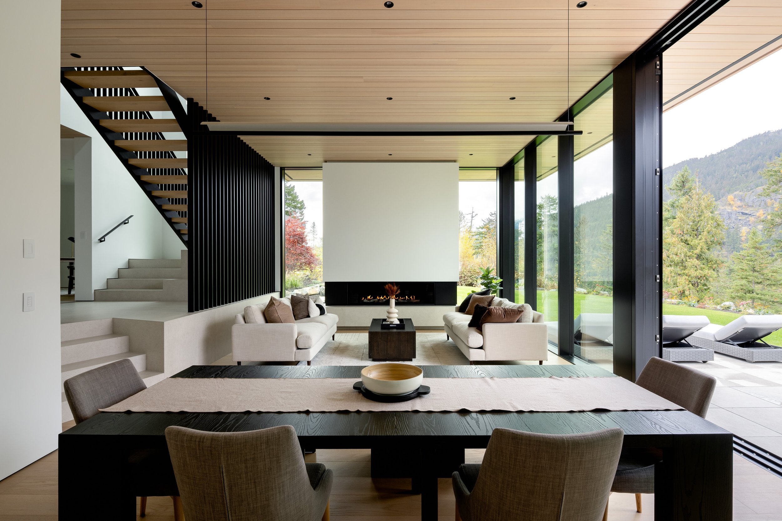 An open-concept interior living and dining space defined by a wood ceiling, large sliding glass walls, and a central fireplace. This image emphasizes spatial flow, natural light, and a strong connection to the outdoor landscape.