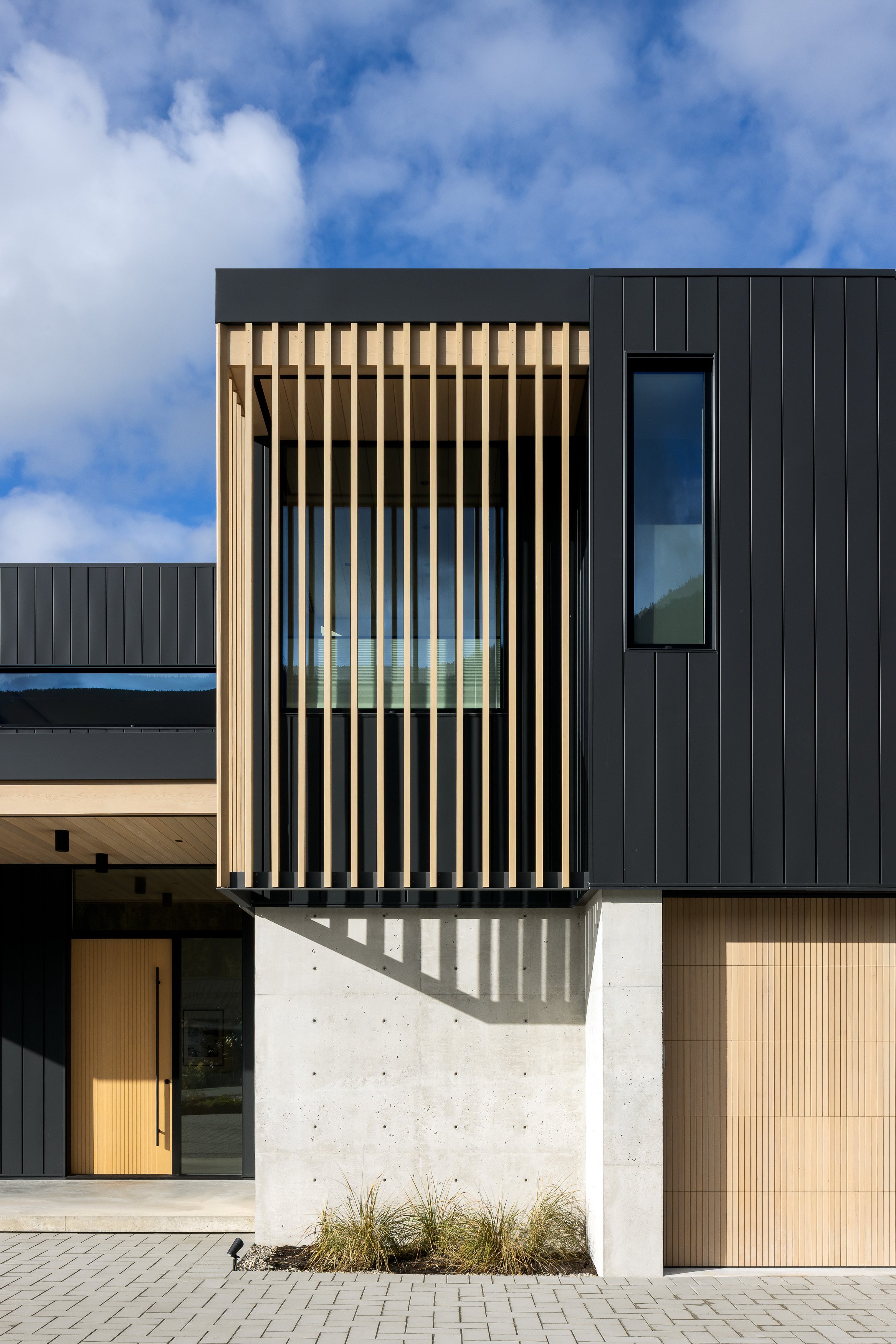 An exterior architectural detail focusing on vertical wood fins, dark cladding, and exposed concrete structure. The composition emphasizes rhythm, shadow, and material depth on the building façade.