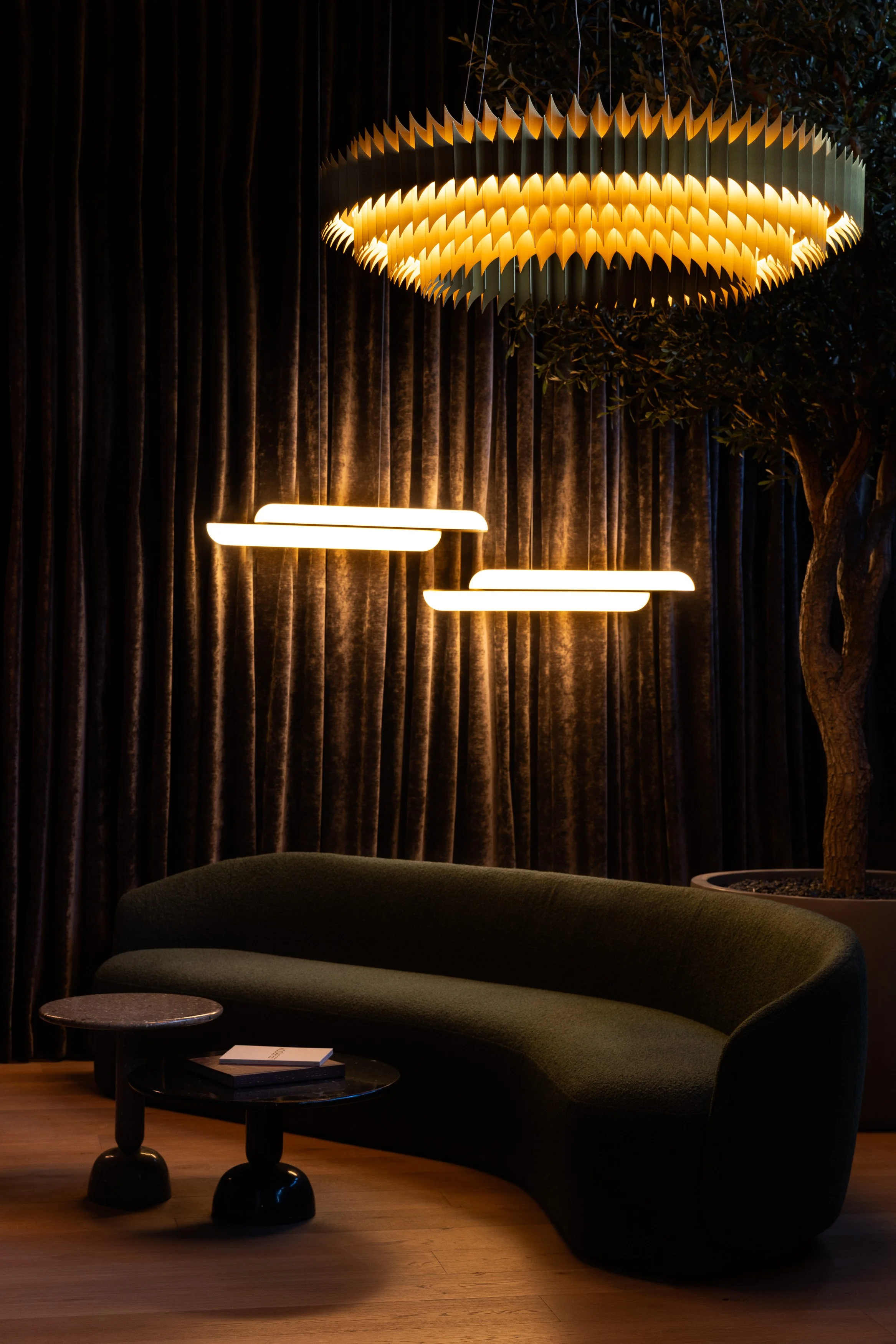Lounge seating area with curved sofa, sculptural chandelier, linear pendants, and dark drapery in the Lightform Vancouver showroom.