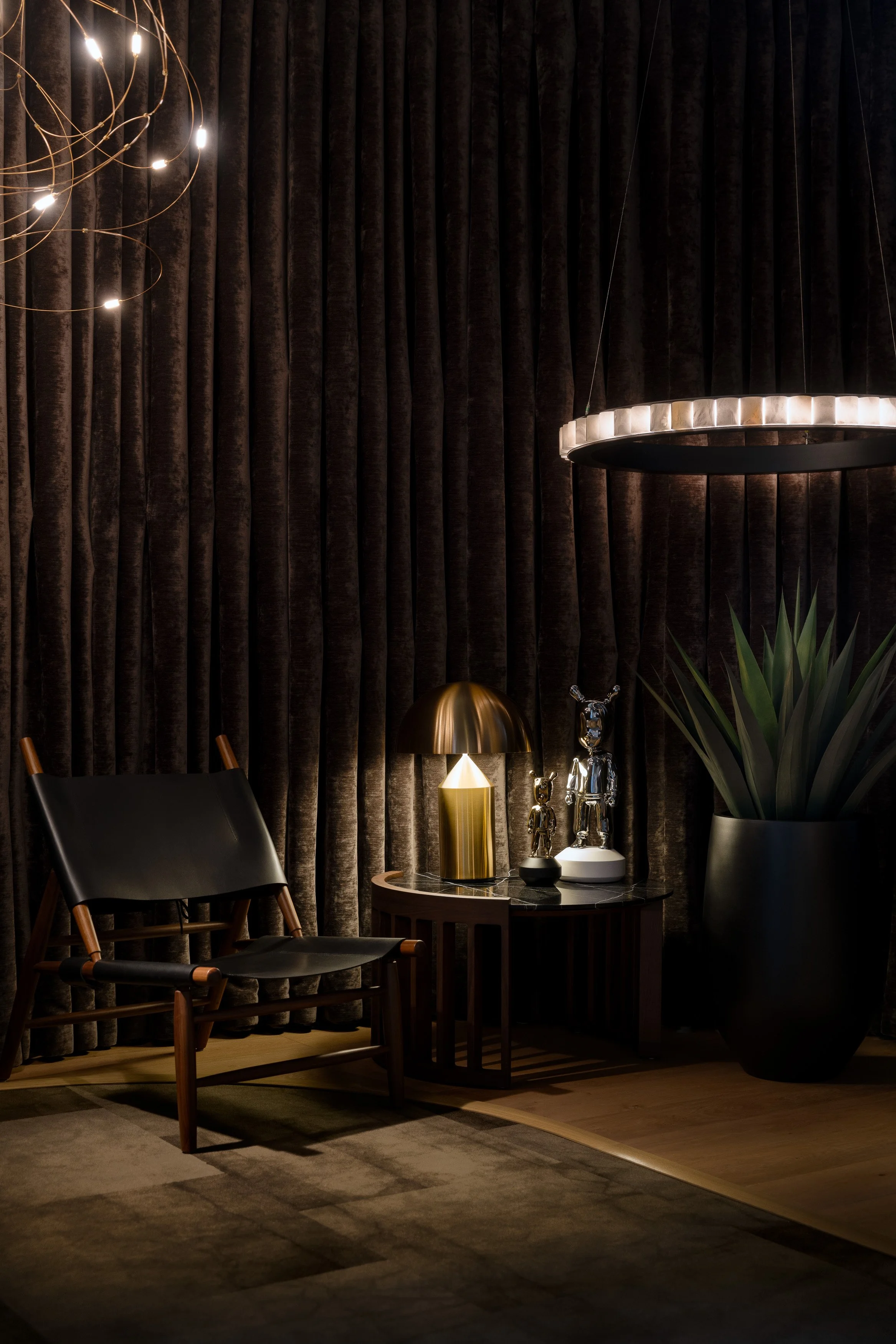 Moody seating vignette with lounge chair, side table lamp, and suspended ring pendant against dark velvet drapery.