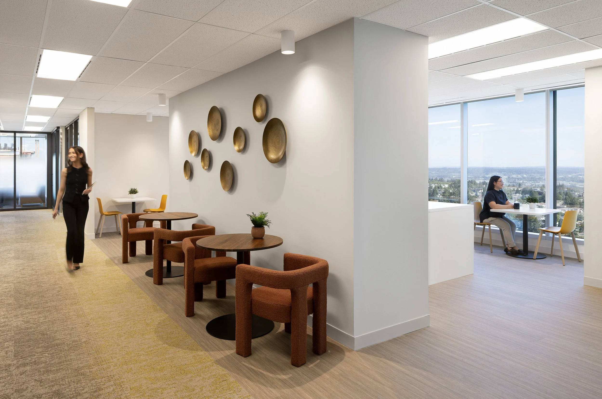 Sun Life office lounge area in Burnaby featuring small round tables, textured lounge chairs, sculptural wall art, floor-to-ceiling windows, and employees using the space.