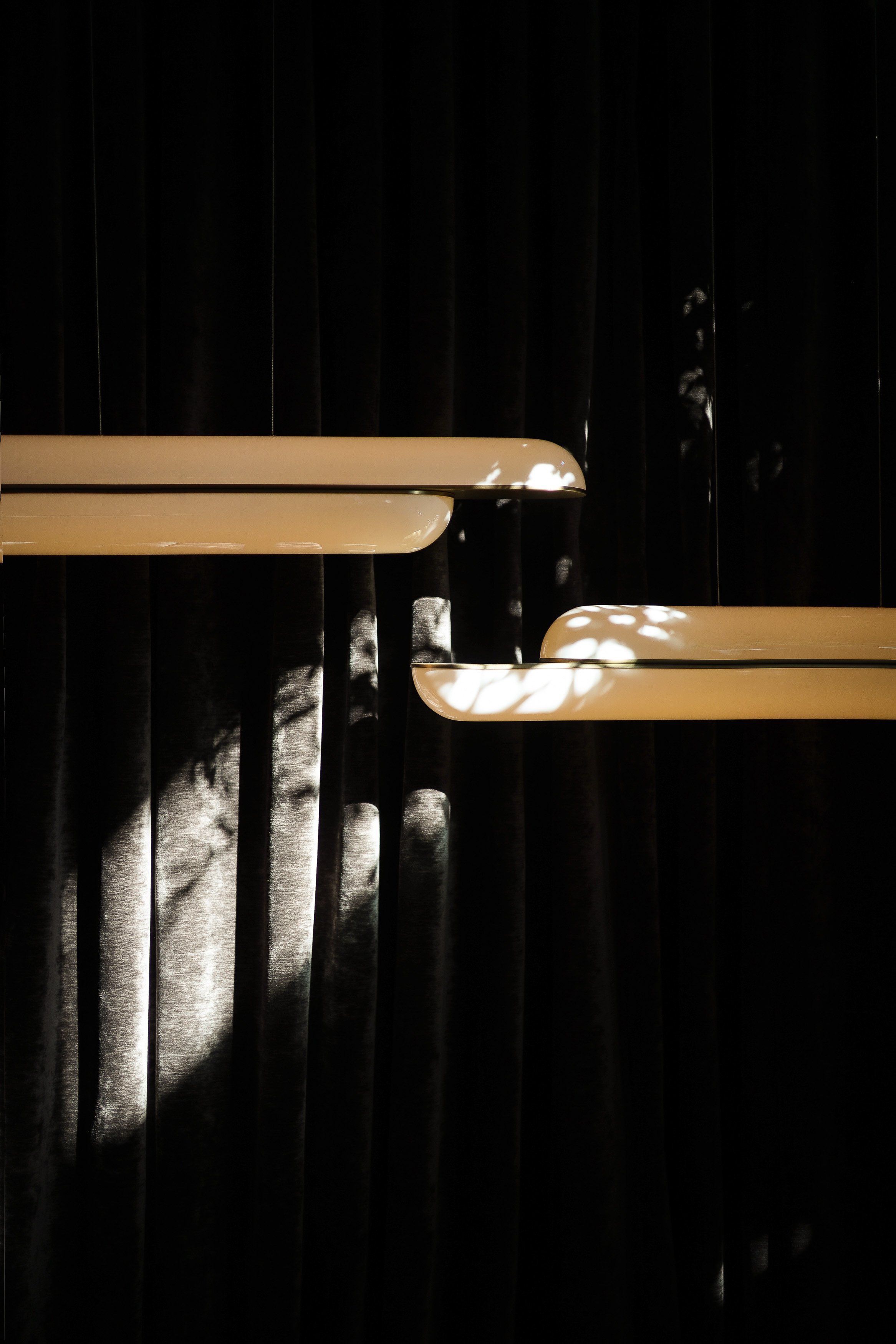 Close-up of two cream linear wall lights mounted in front of a dark velvet curtain with dappled daylight highlights.