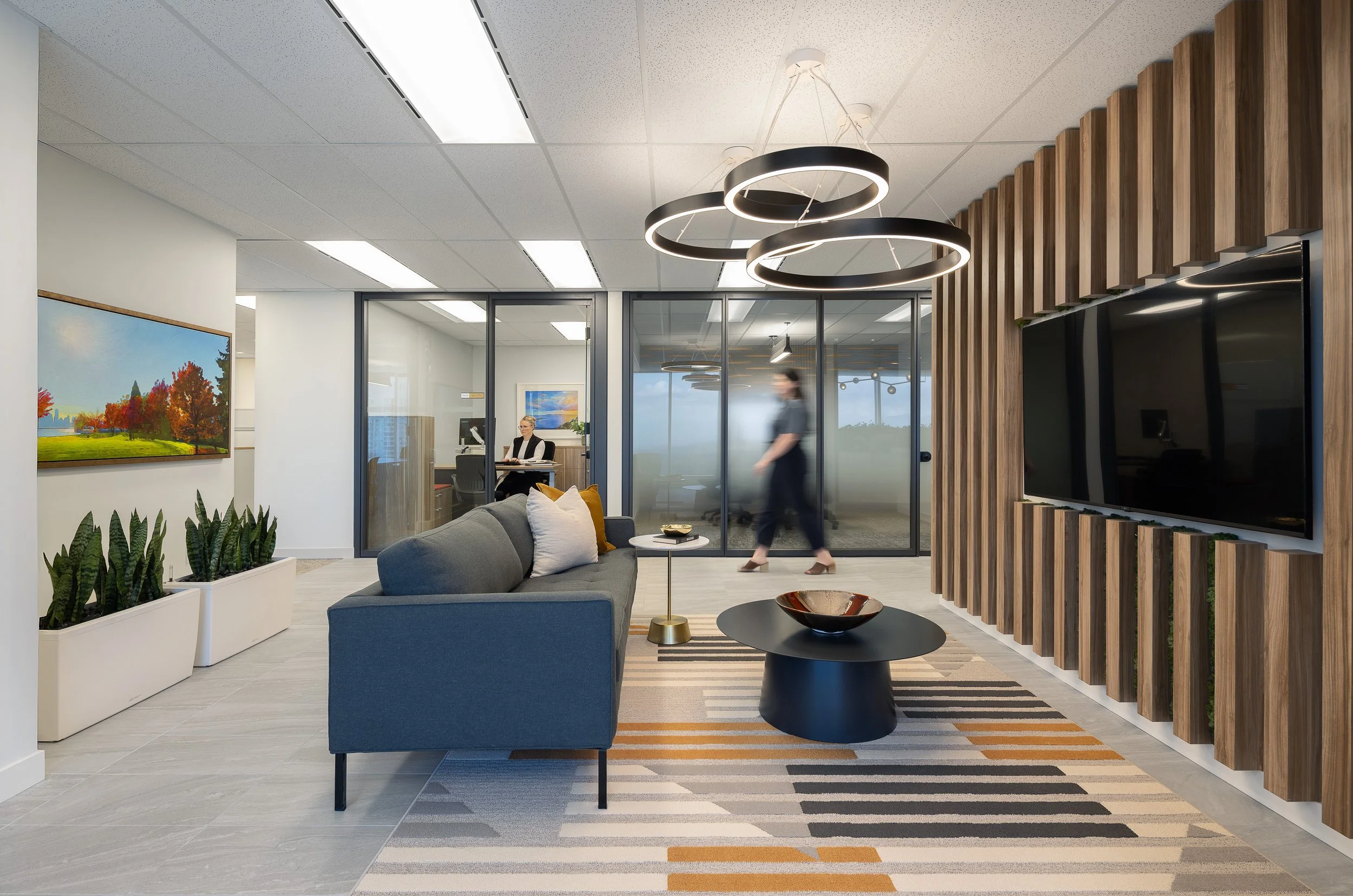 Sun Life office lounge area in Burnaby with a sofa, coffee table, wall-mounted screen, wood feature wall, and glass-walled meeting rooms in the background.