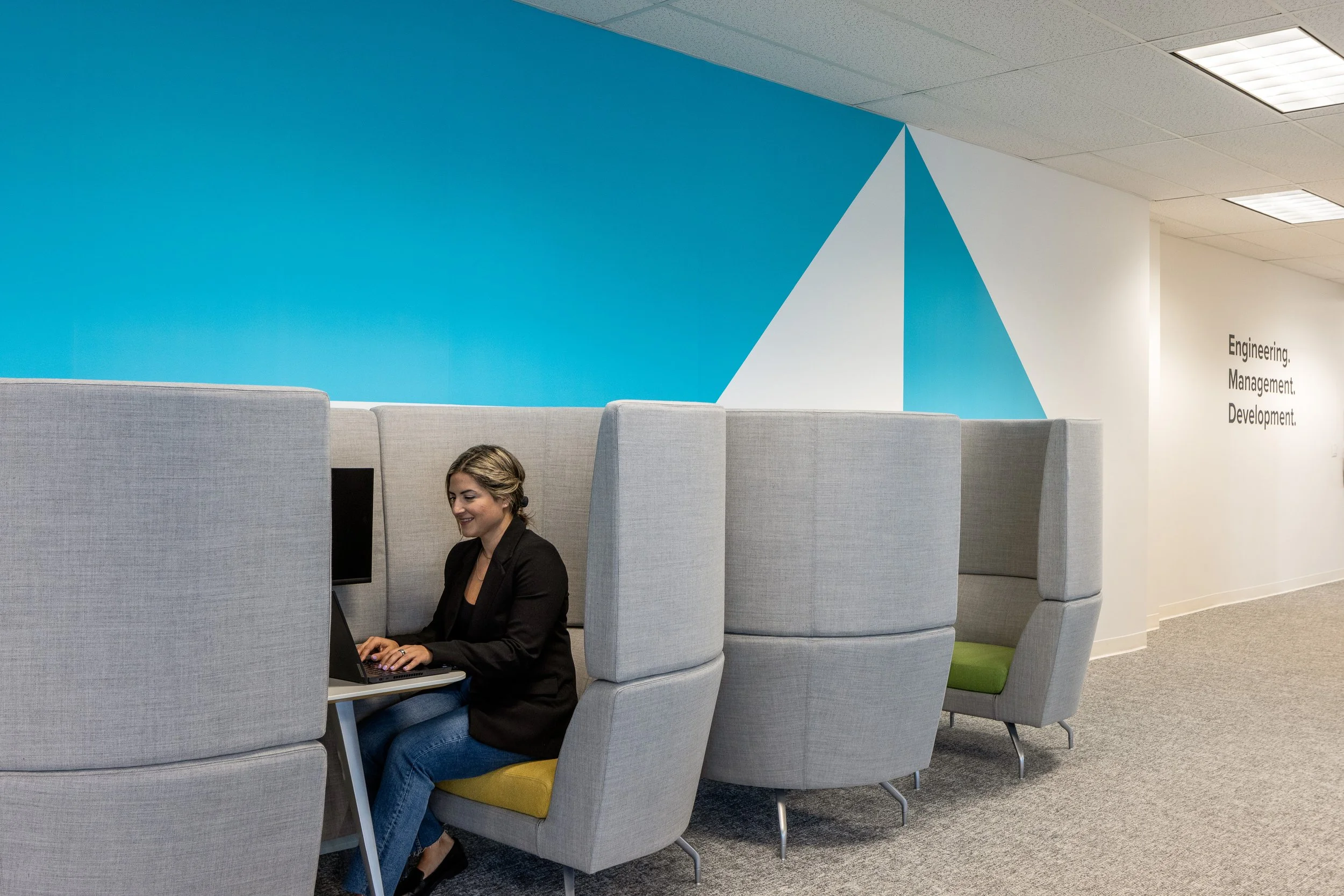 Acoustic work booths along a turquoise feature wall at the Mott MacDonald Vancouver office, with employee working on a laptop.