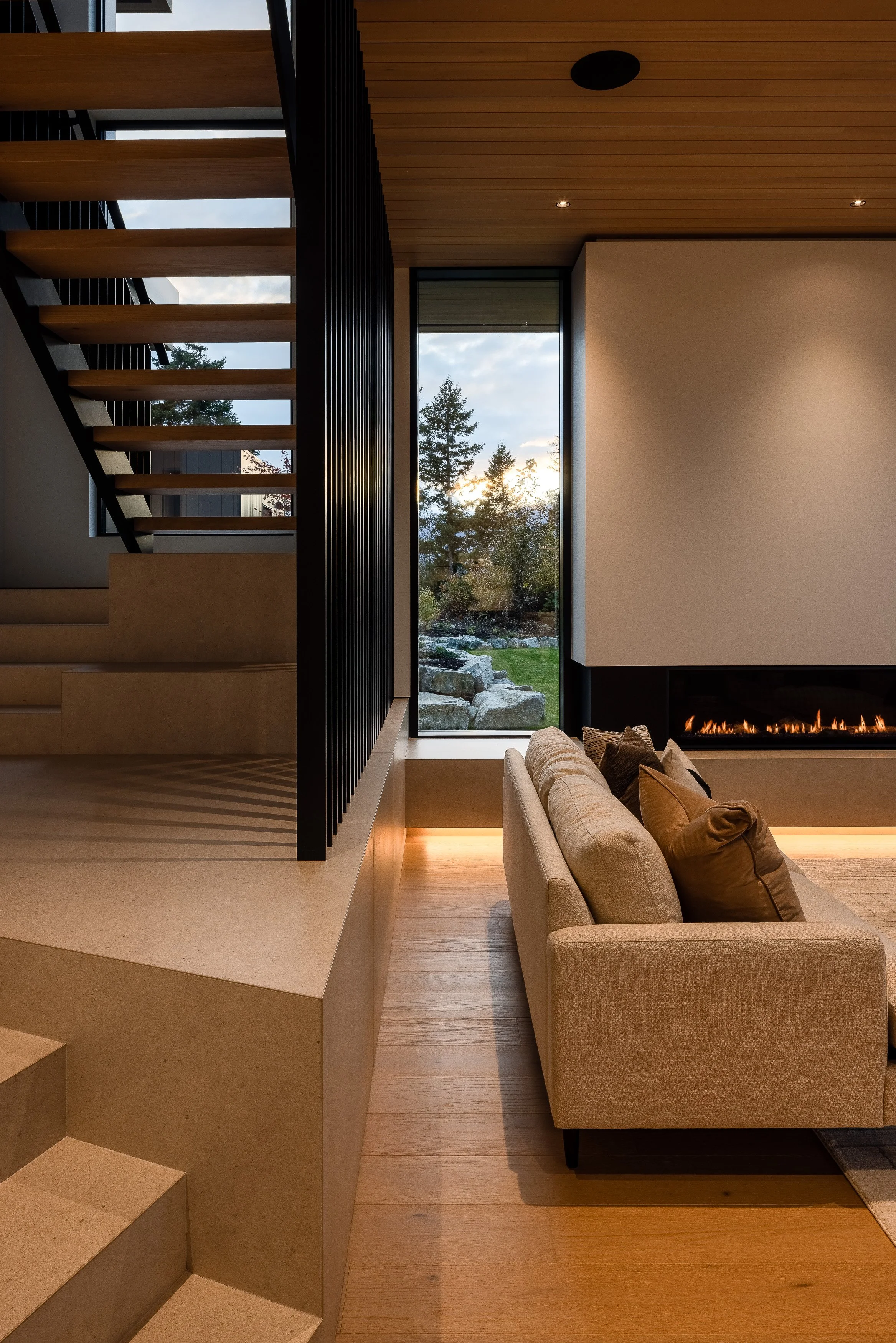 Interior dusk: stair + fireplace wall, tall window to landscape, warm interior glow.