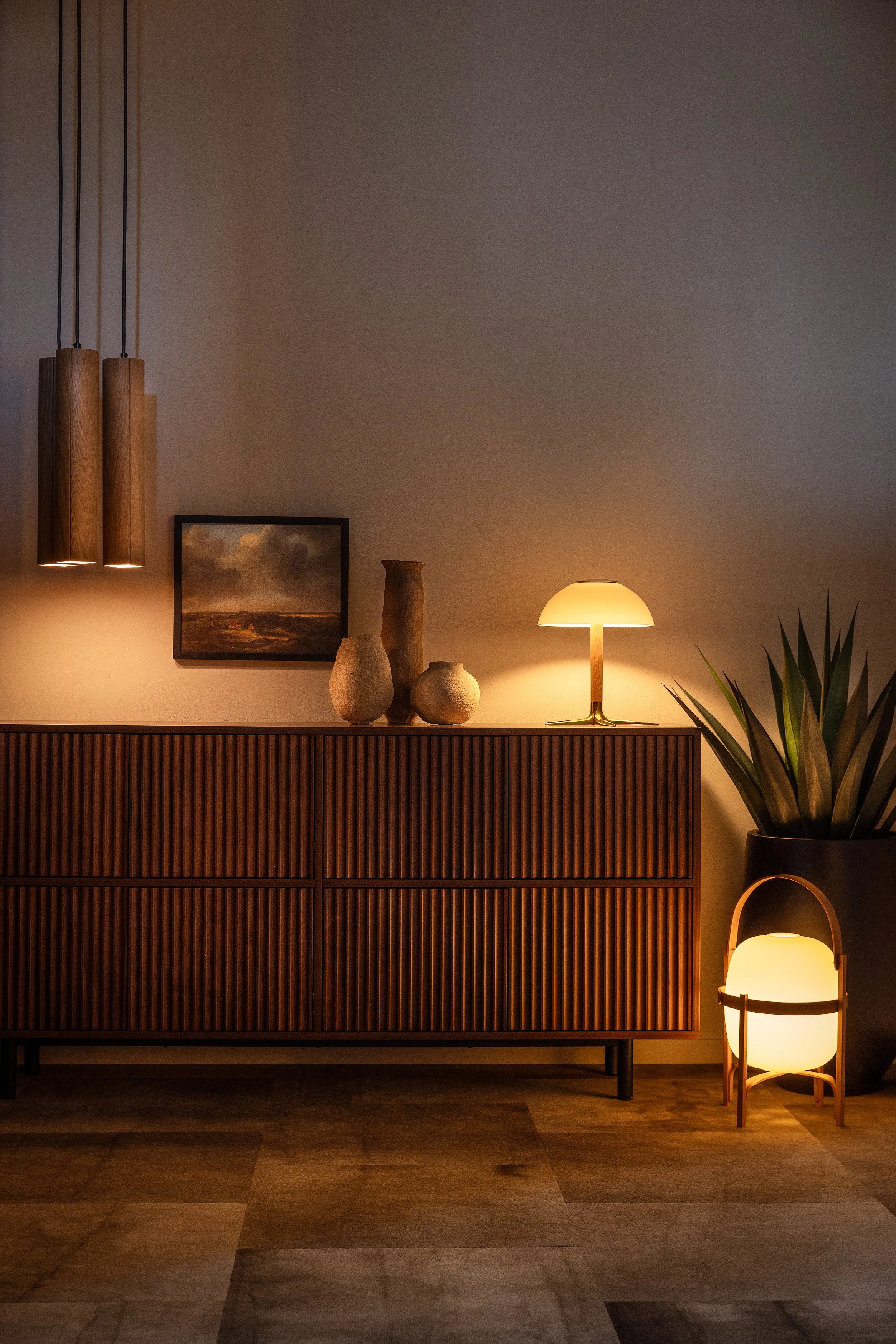 Wide showroom vignette with ribbed credenza, paired wood cylinder pendants, a dome table lamp, ceramics, and a floor lantern.
