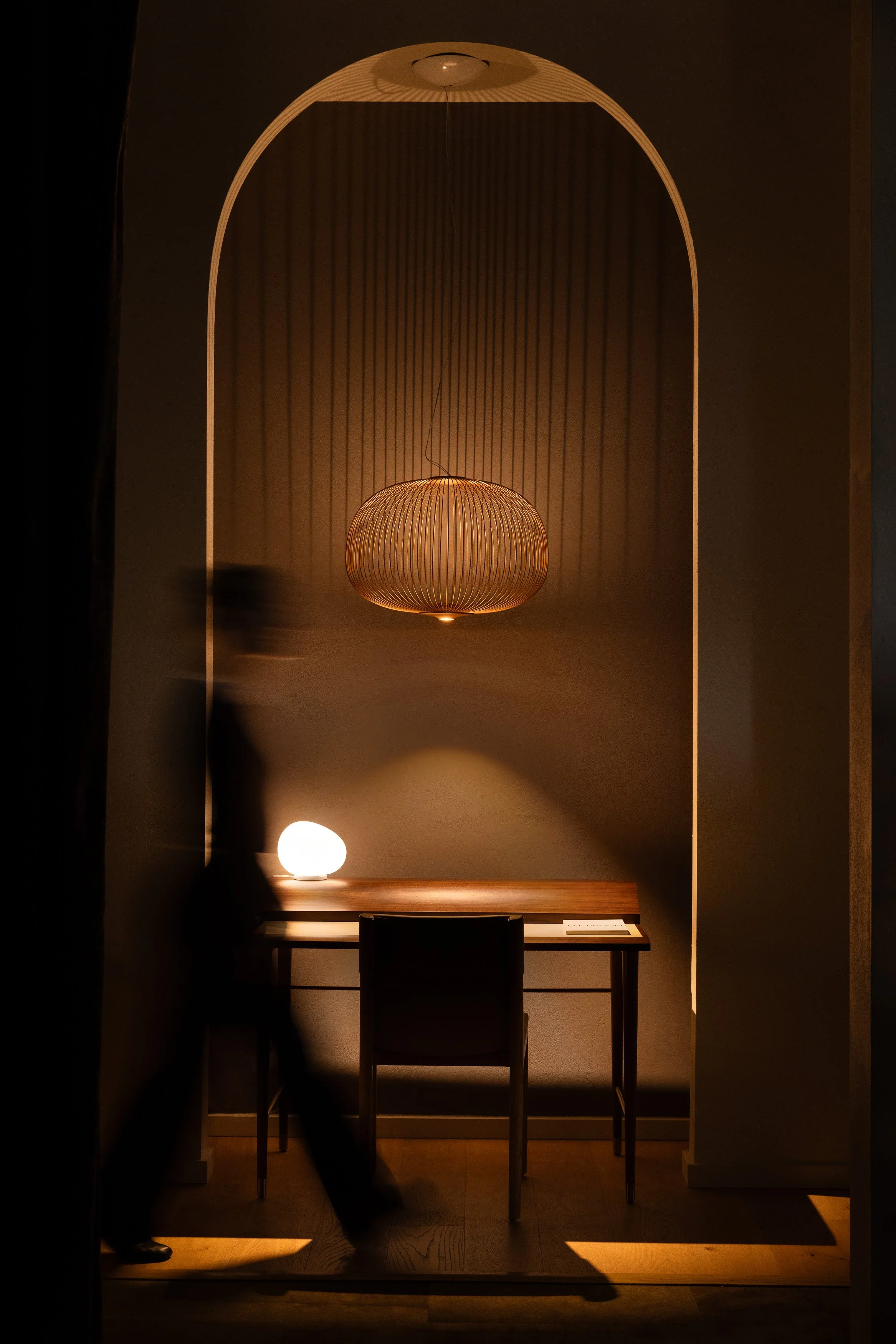 Arched alcove workspace with desk, chair, woven pendant light, and a blurred figure passing through warm light.