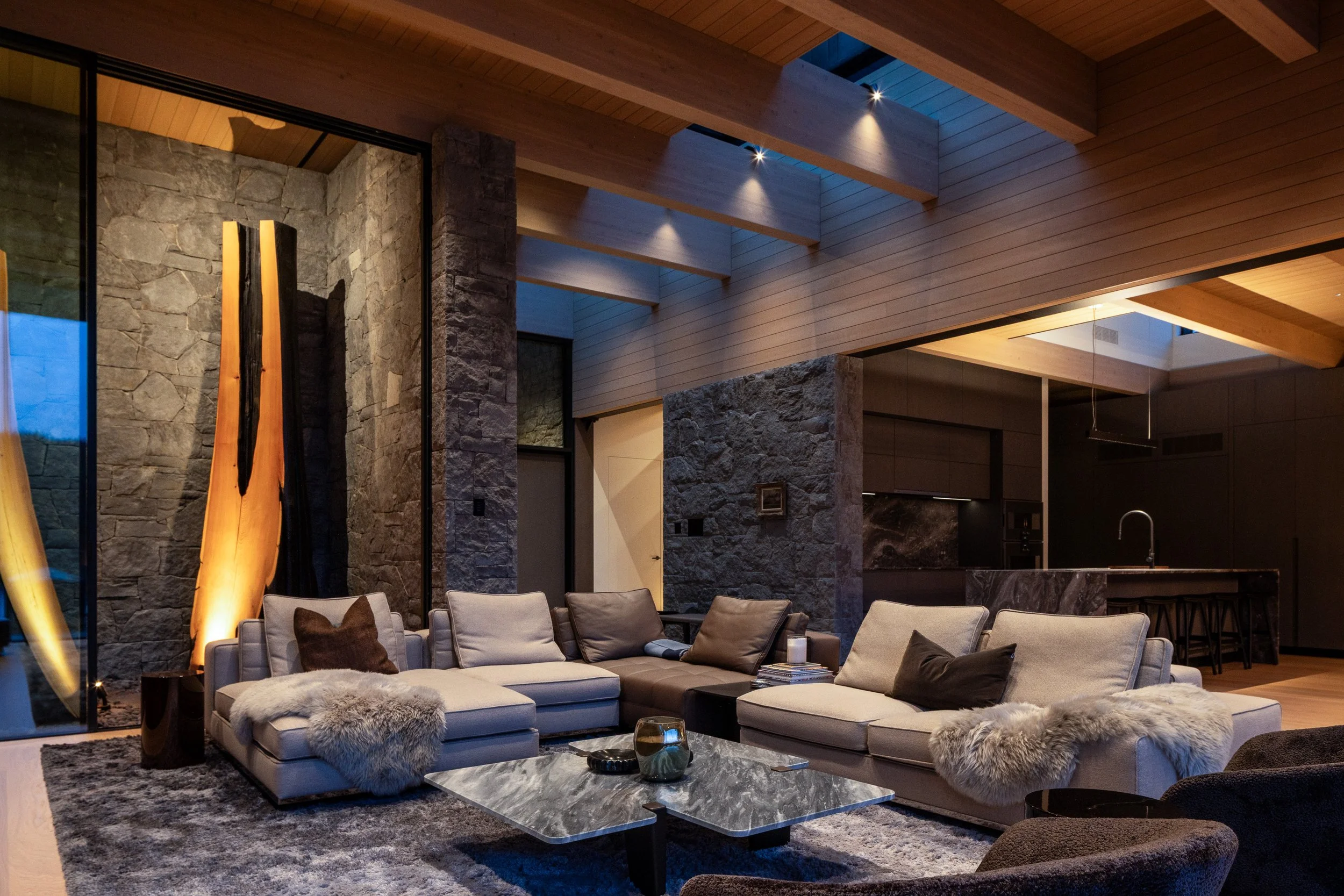 Modern living room with beige sectional sofa, fur throws, and dark pillows, featuring stone walls, wooden ceiling beams, and an open kitchen in the background.