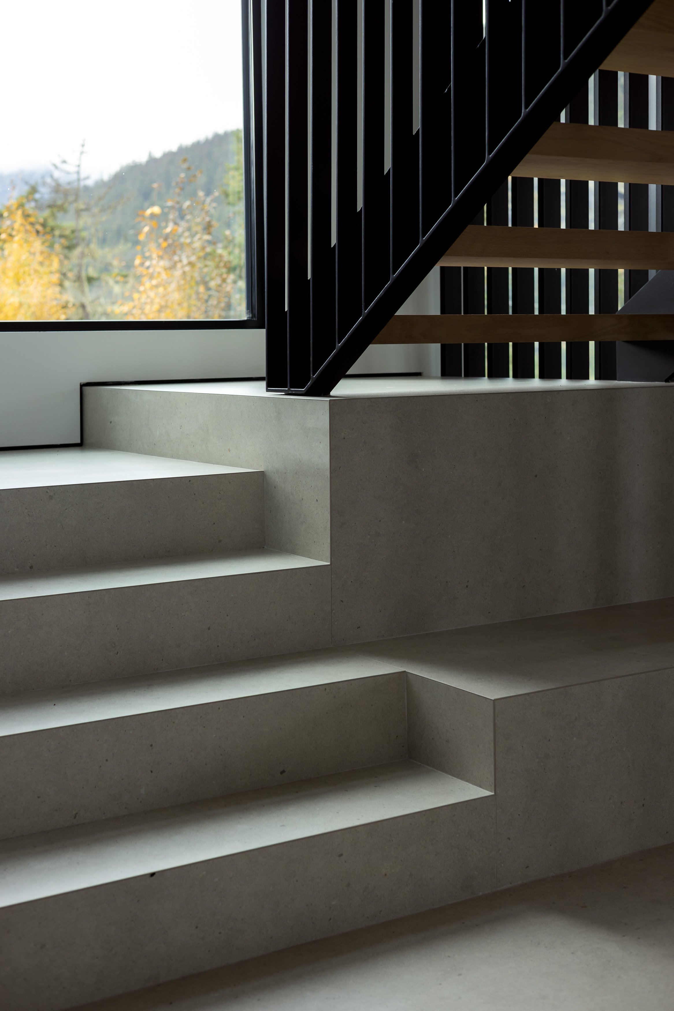 A detailed interior view of the stair and landing, featuring board-formed concrete, open wood treads, and vertical steel balustrades. The image highlights precision detailing and material transitions within the home.