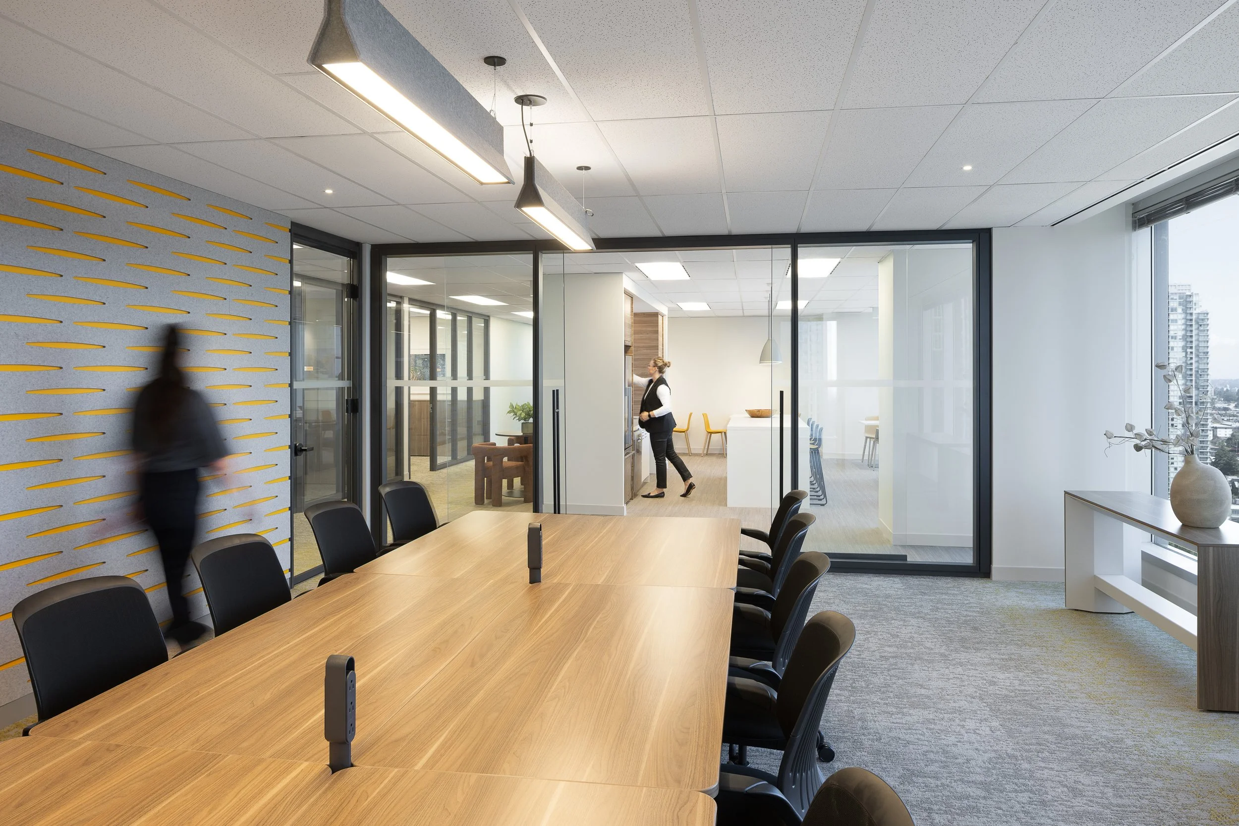 Sun Life office meeting room in Burnaby with a long wood conference table, black chairs, glass-walled rooms, patterned acoustic wall panels, and staff moving through the space.