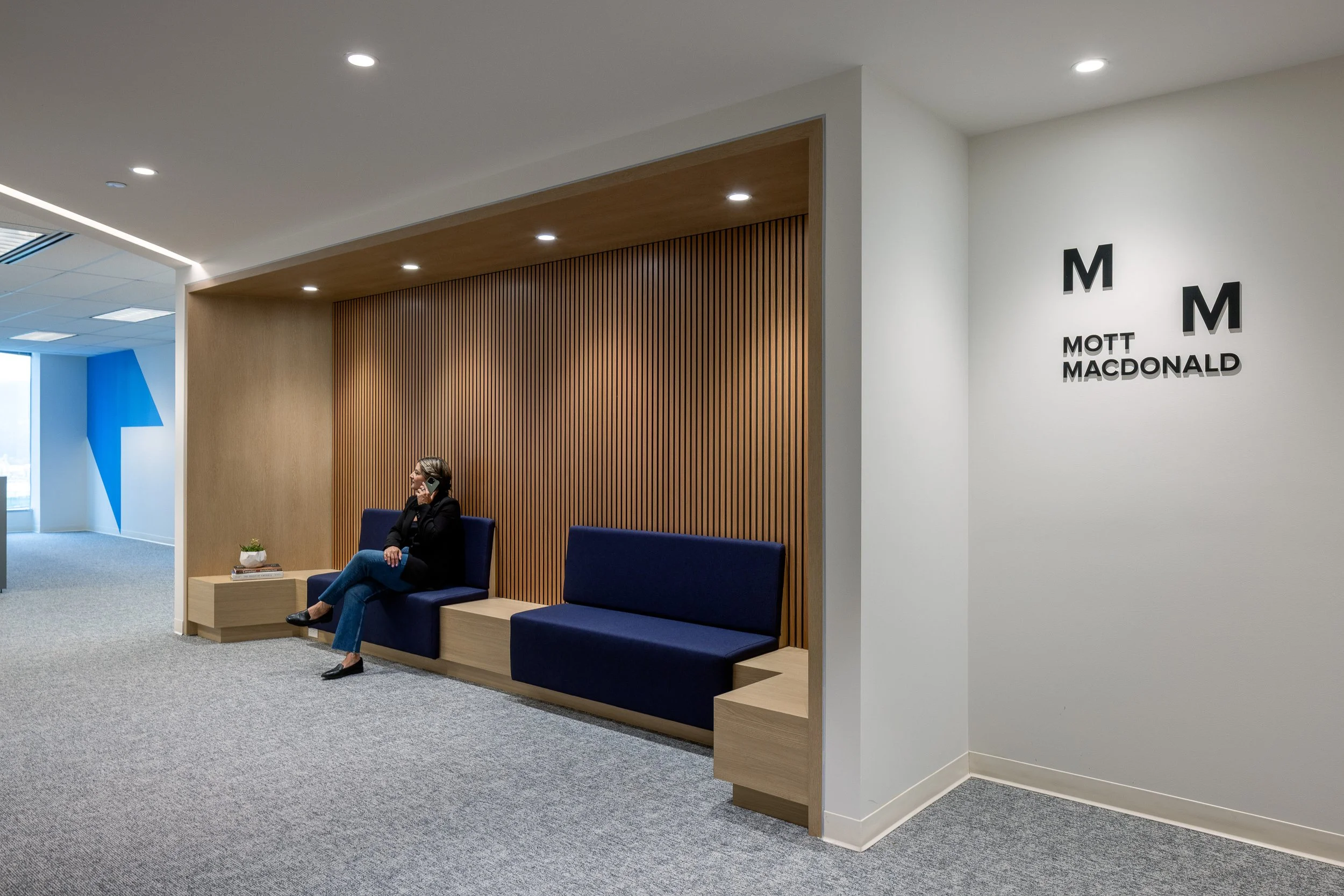 Built-in lounge seating with wood slat feature wall at the Mott MacDonald Vancouver office, with employee seated beneath the company logo.