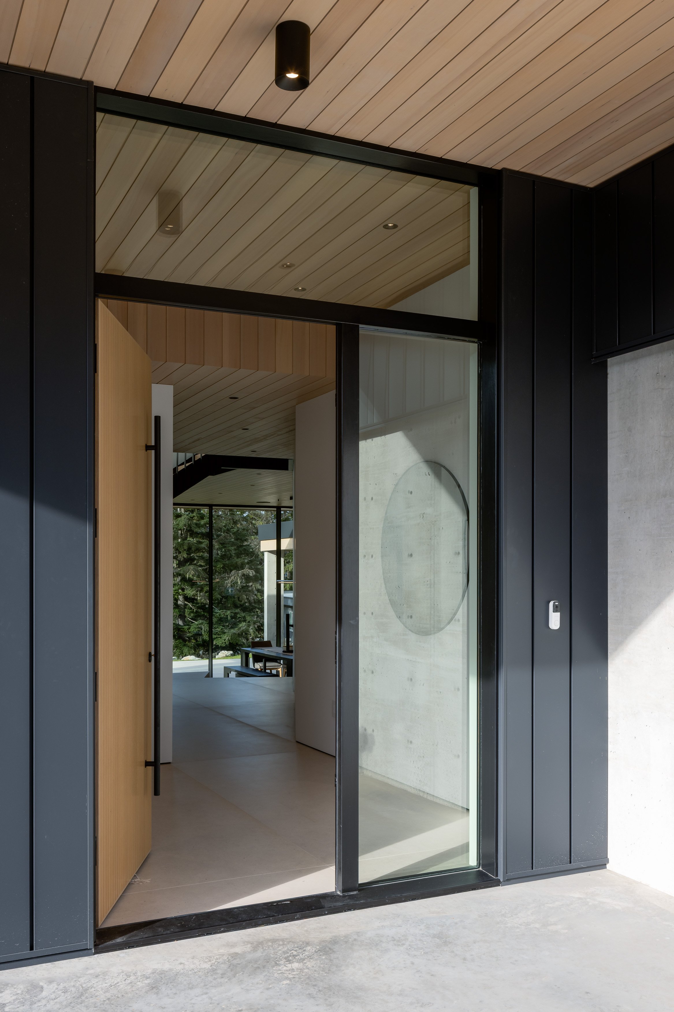 Entry detail: open wood front door, tall glazing/sidelight, black cladding + concrete wall.