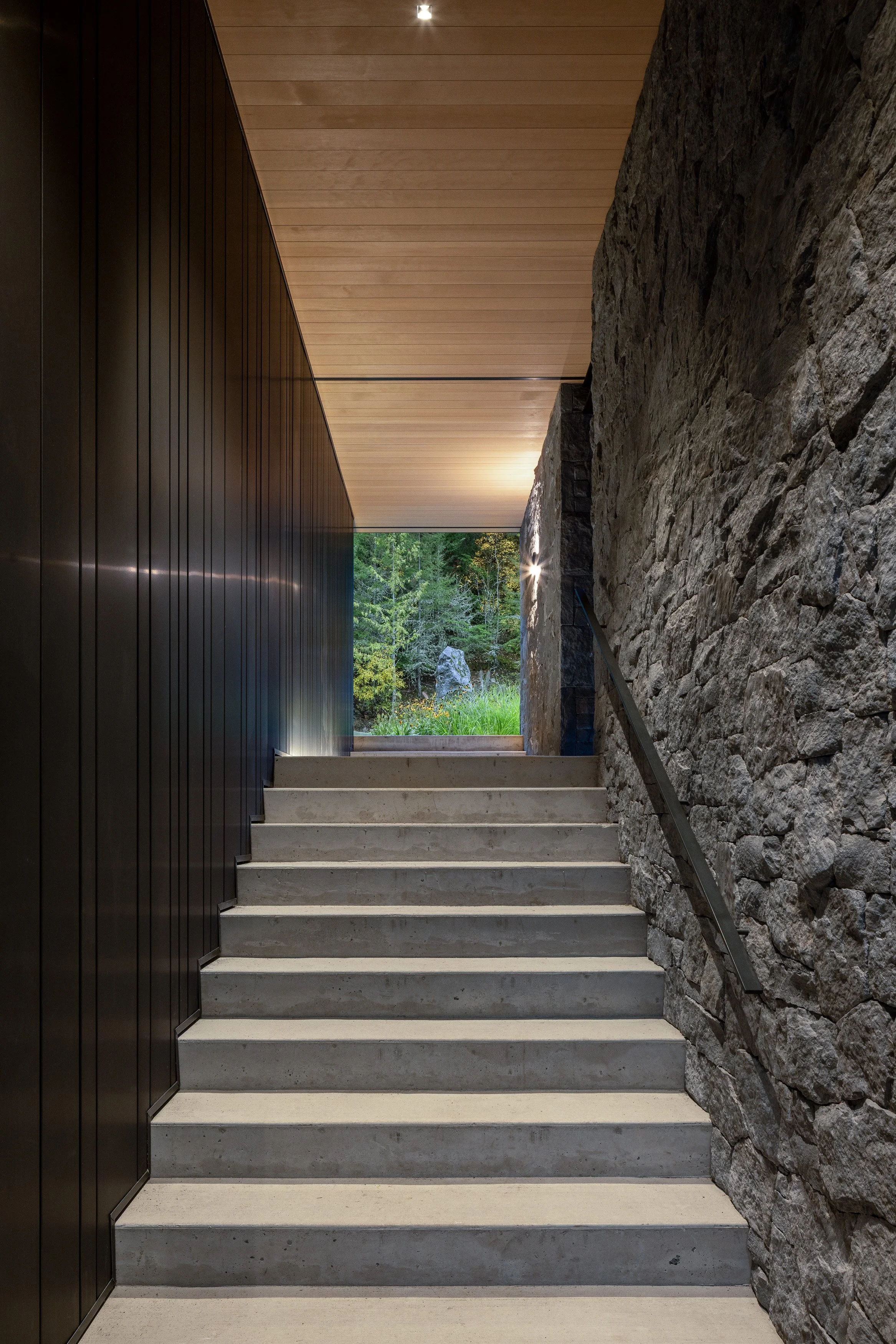 A dramatic interior stairway defined by board-formed concrete steps, dark vertical cladding, and a natural stone wall. Designed by Burgers Architecture and built by Durfeld Constructors, this Whistler home uses material contrast and controlled light 