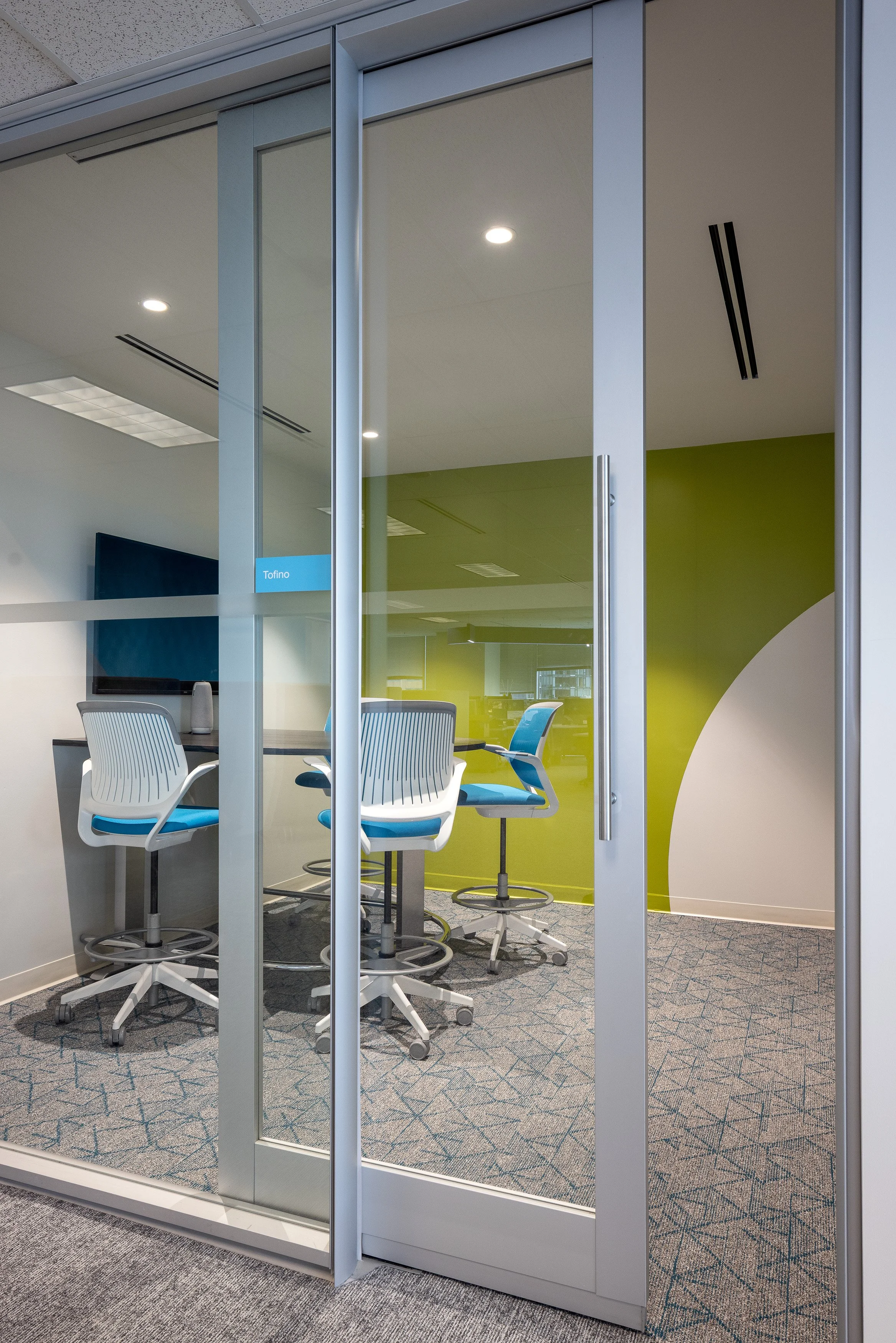 Glass-enclosed collaboration room with high stools and green accent wall at the Mott MacDonald Vancouver office, designed by Aura Office Interiors.