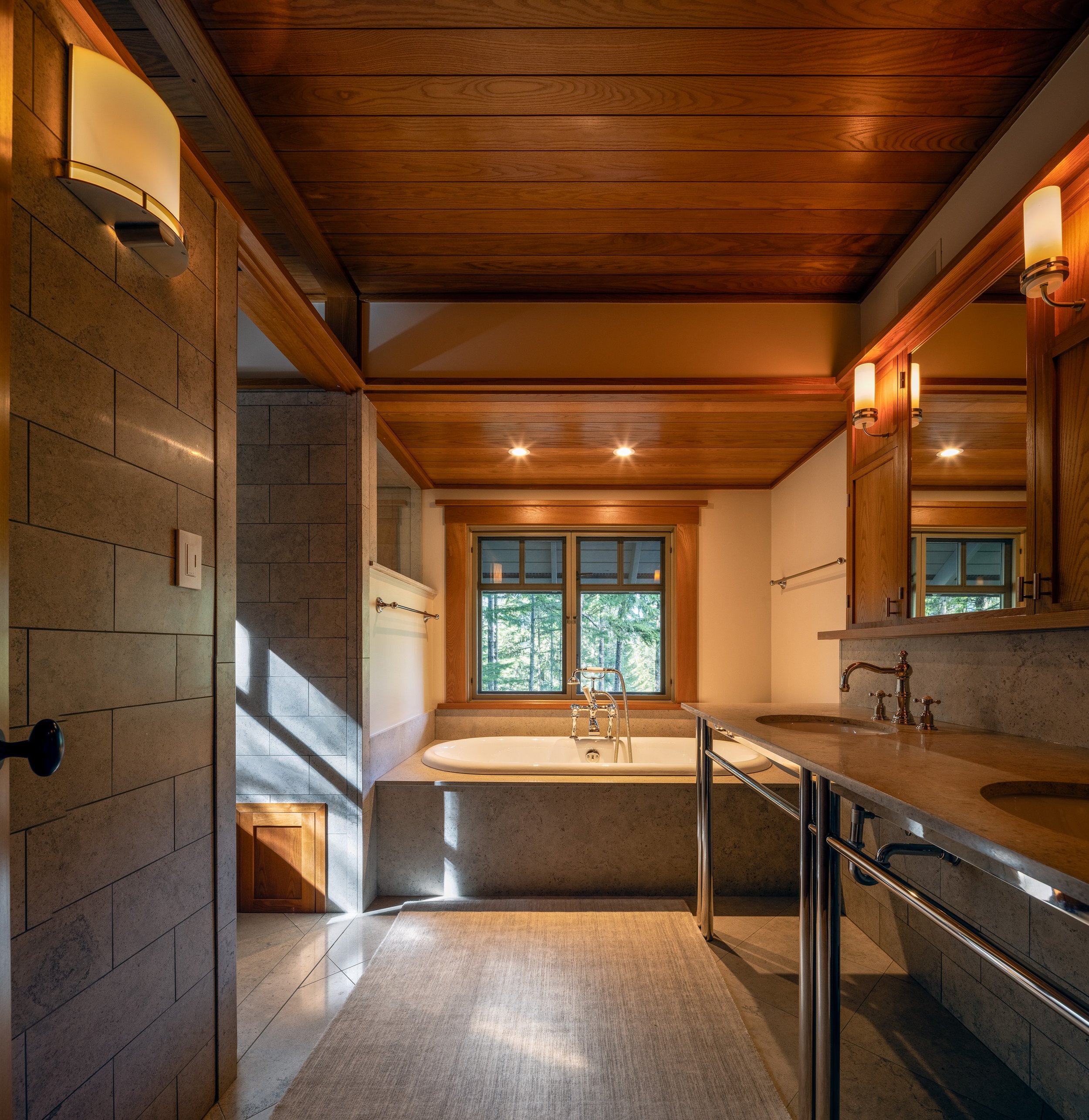 Bathroom with stone tile, wood ceiling, double vanity, and a deep soaking tub with a forest-facing window in Whistler.