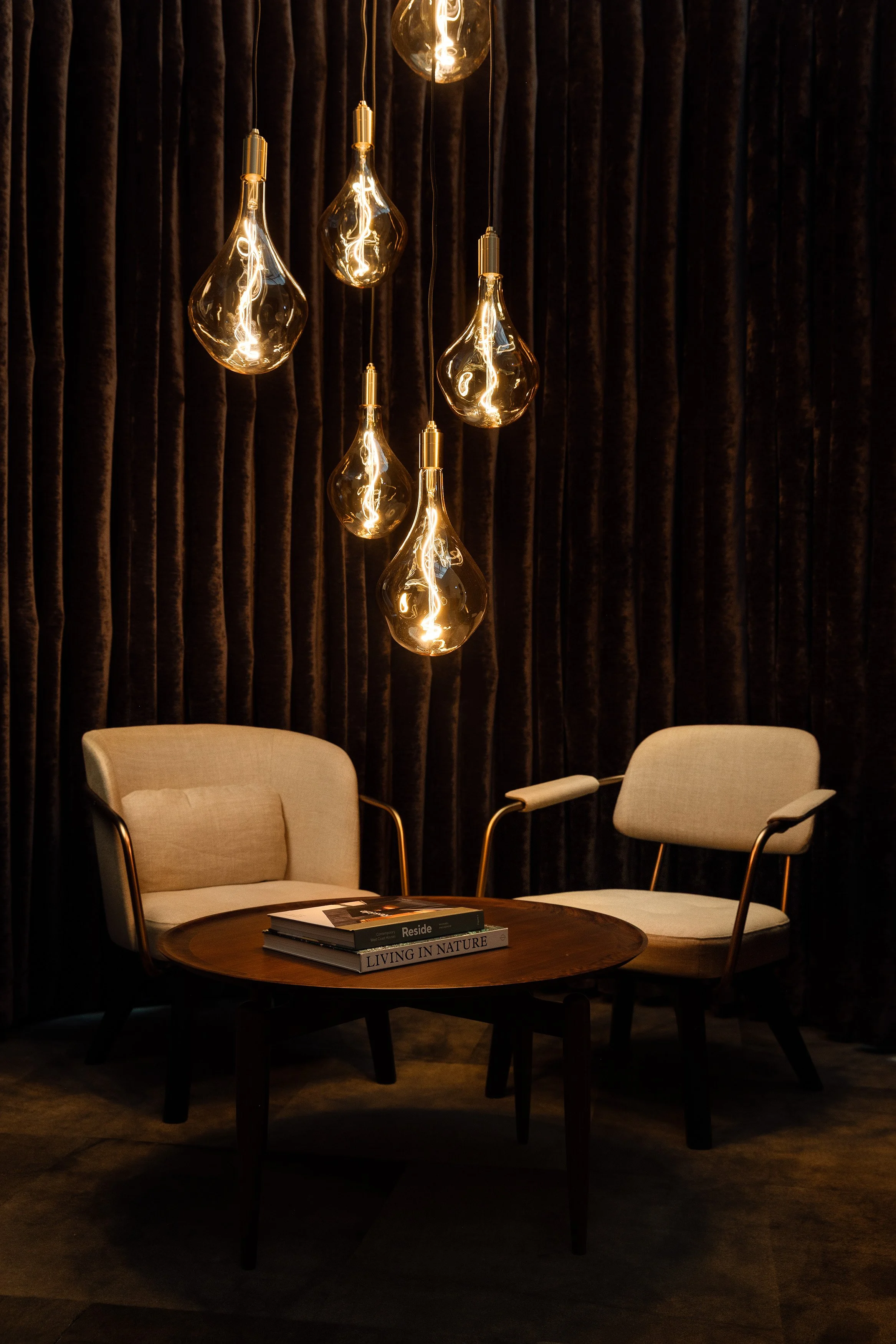 Two lounge chairs and round table beneath oversized teardrop glass pendants, set against dark velvet curtains.