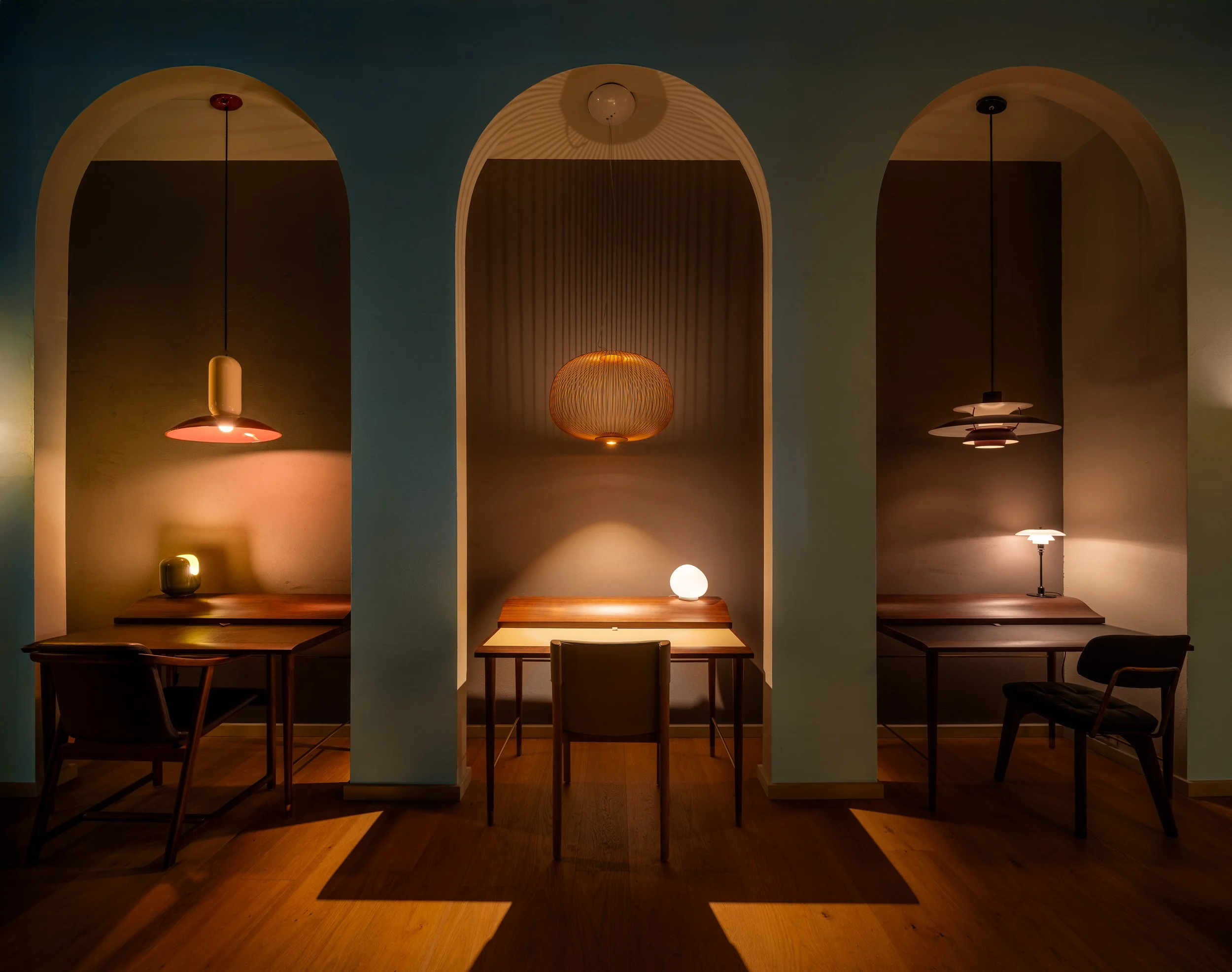 Three individual work spaces separated by white arches, each with a desk, chair, and distinct light fixture, creating a warm and cozy atmosphere.