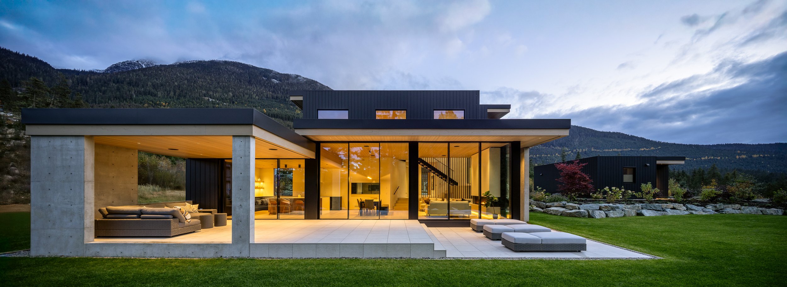 Exterior dusk hero (panoramic): house fully lit, big glazing, patio + loungers, mountains behind.