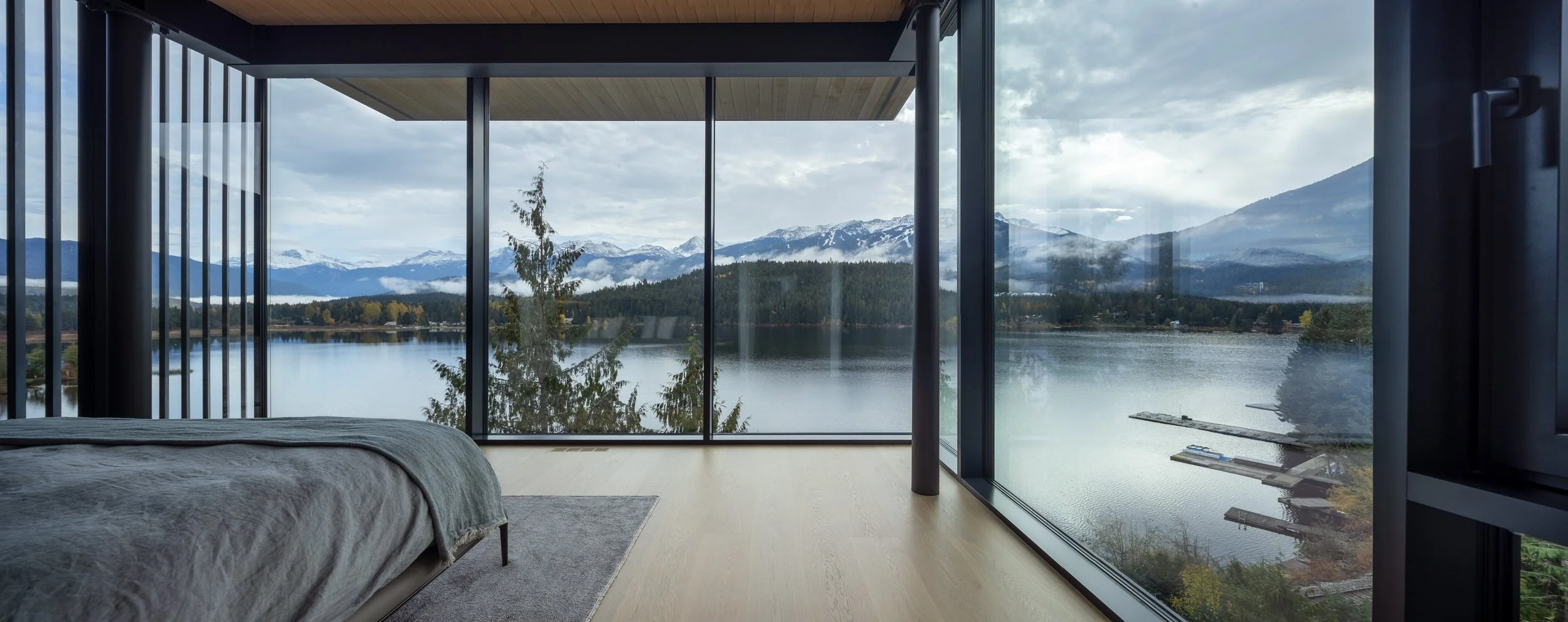 A serene Whistler bedroom with expansive corner glazing overlooking the lake and surrounding mountains. Built by Durfeld Constructors and designed by Burgers Architecture, the space prioritizes natural light, views, and a calm, understated interior a