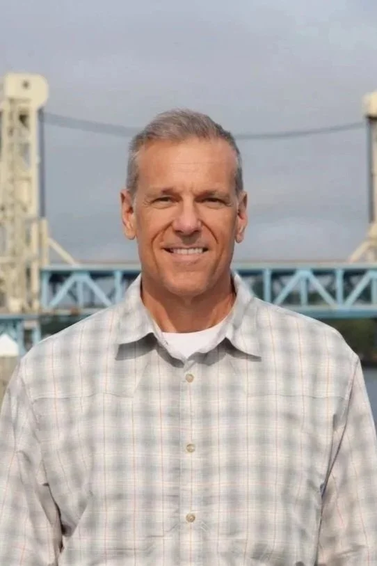 Smiling man in a dark collared shirt in front of a brick building.