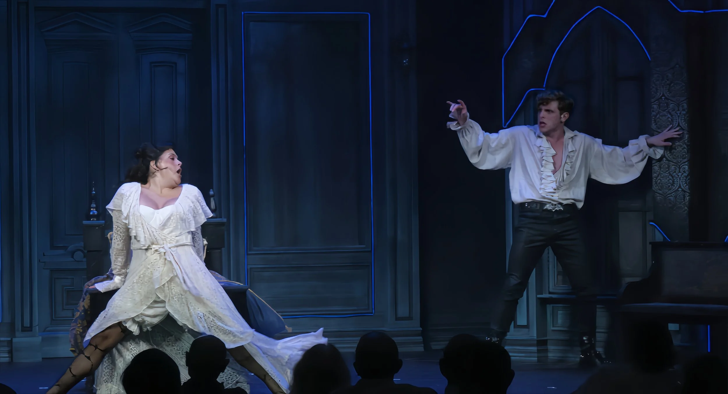 Actors performing a theatrical scene on stage with dark blue background, two large dark wooden panels, and stylized neon lighting.