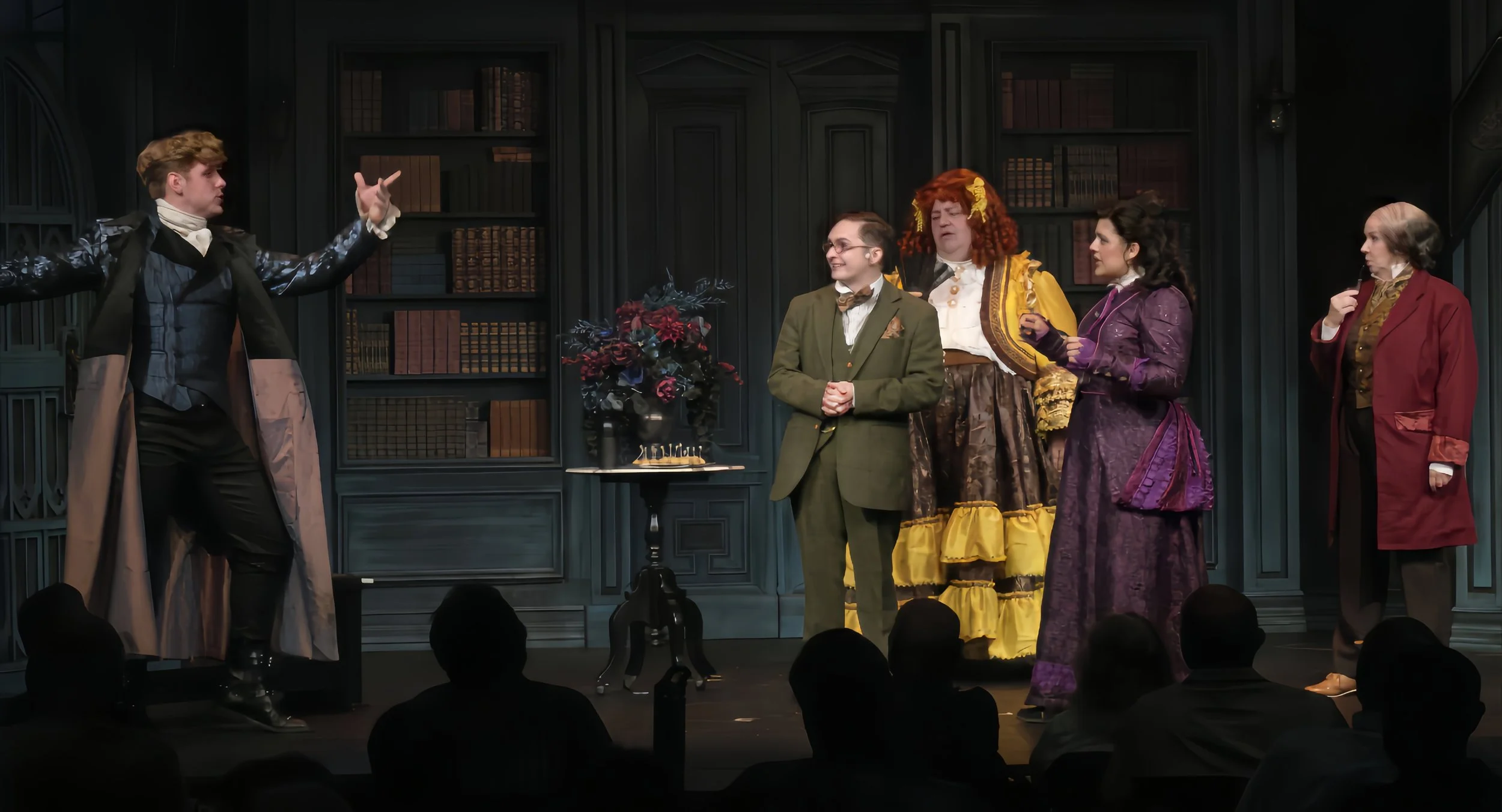 Actors in costume performing on stage in front of an audience, with a dark green bookcase backdrop and a small table with a floral arrangement.