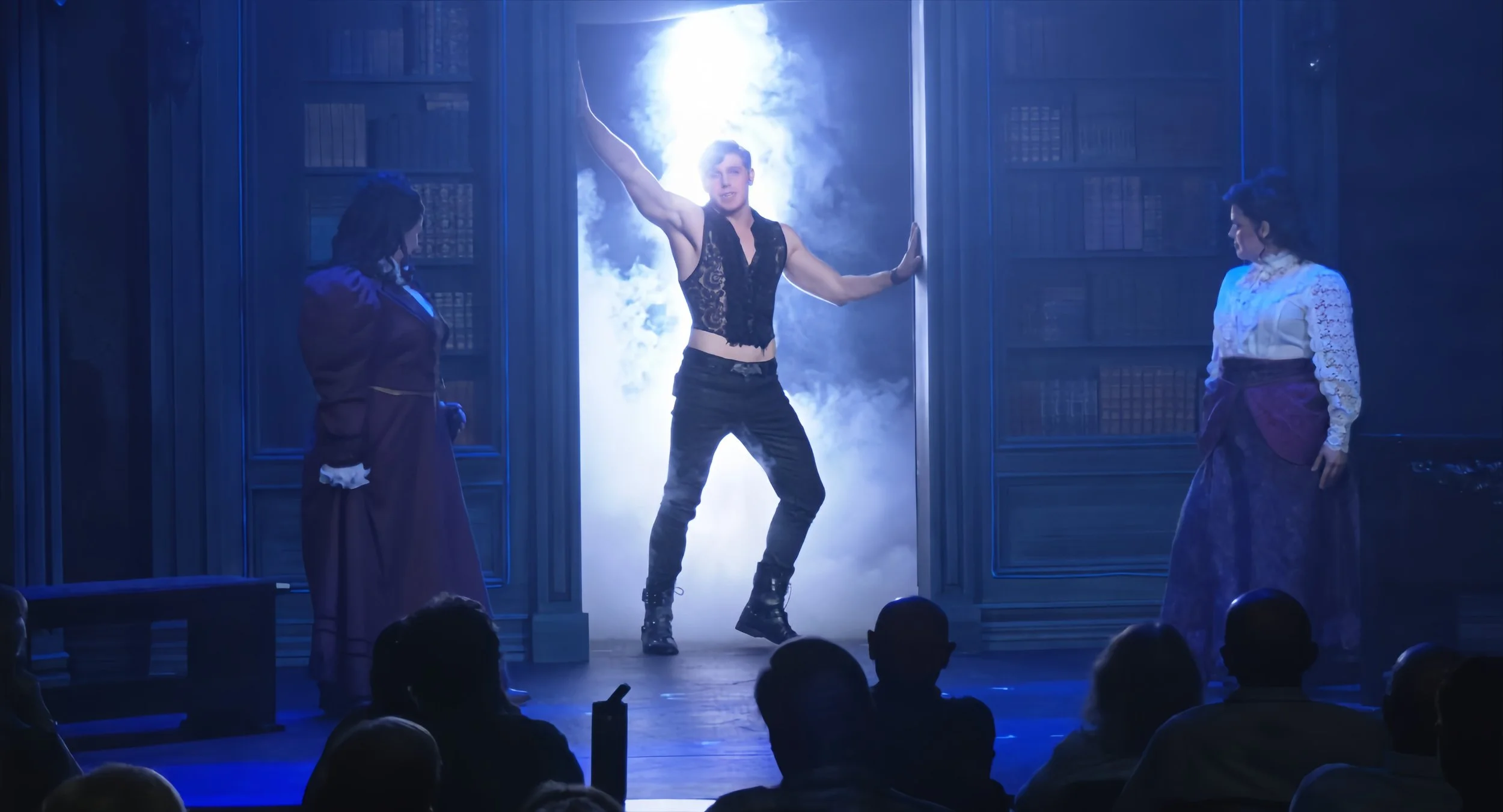 A male performer poses dramatically on stage, with a blue-illuminated foggy background, flanked by two women in vintage dresses watching him.