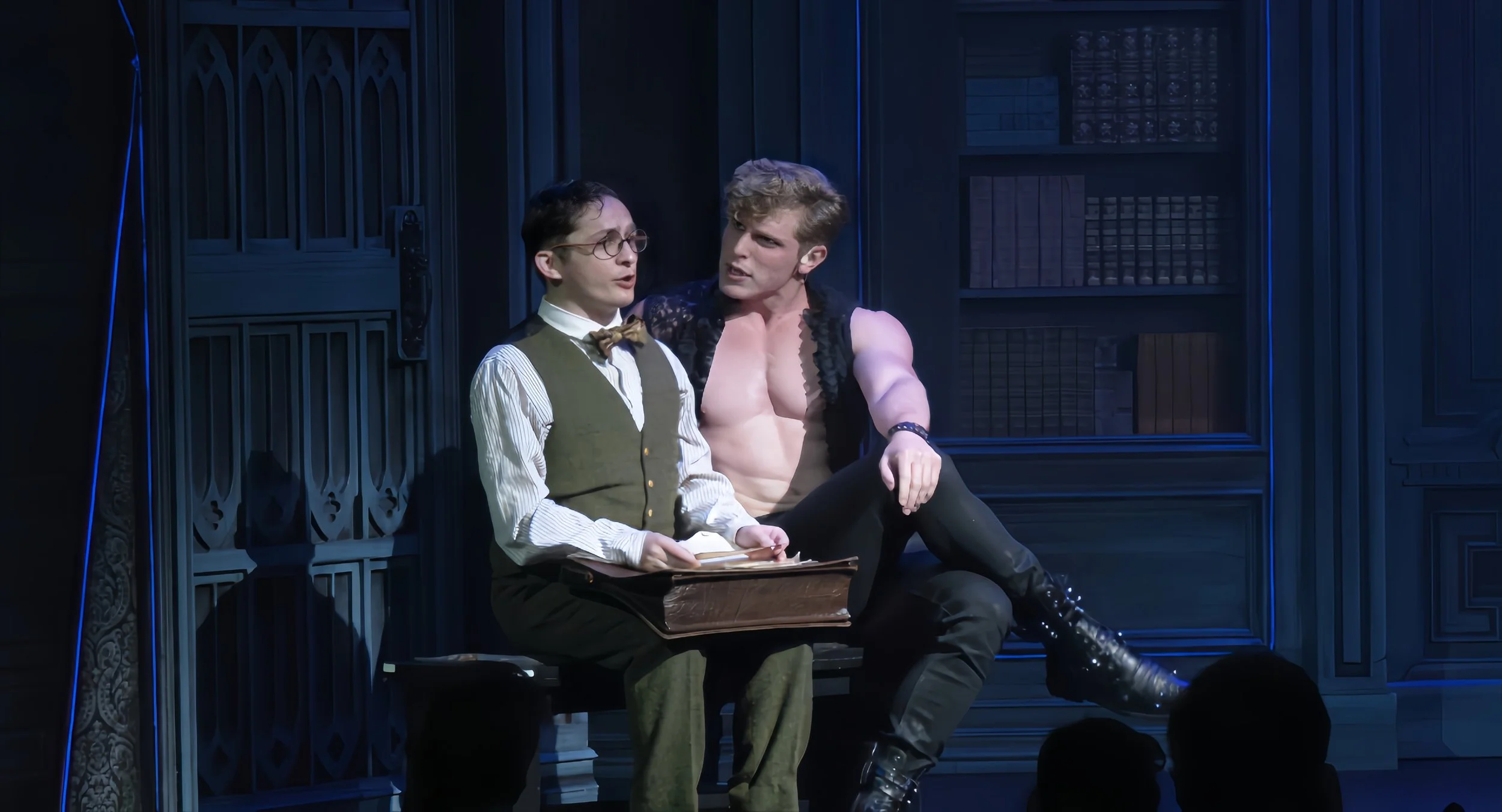 Scene from a play or performance showing two actors on stage. One actor in vintage clothing with glasses and a bow tie, holding a book, looks surprised or confused. The other actor, shirtless with muscular arms, black boots, and leather straps, is si