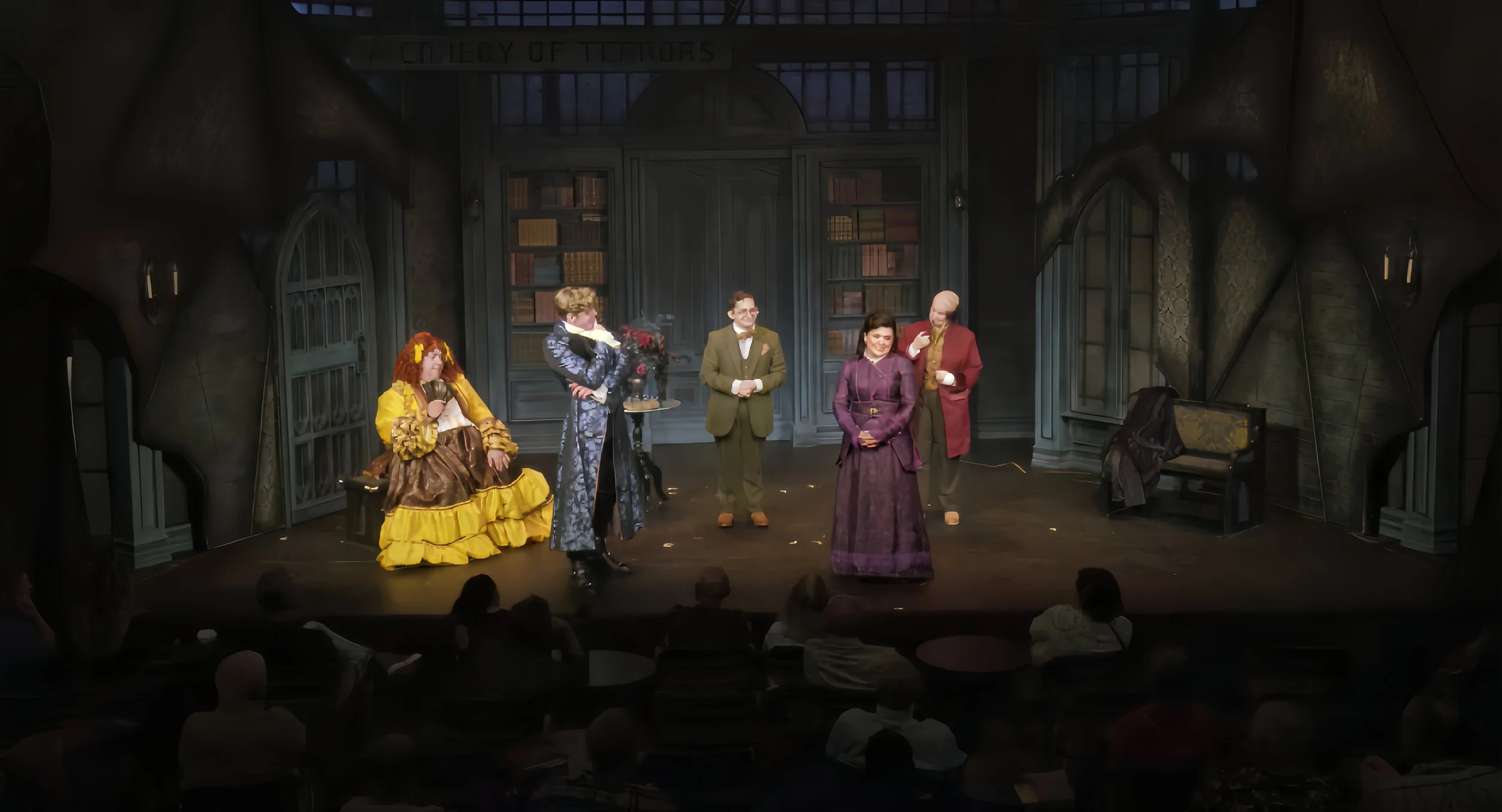 Theater stage with six actors in period costumes performing, audience watching.