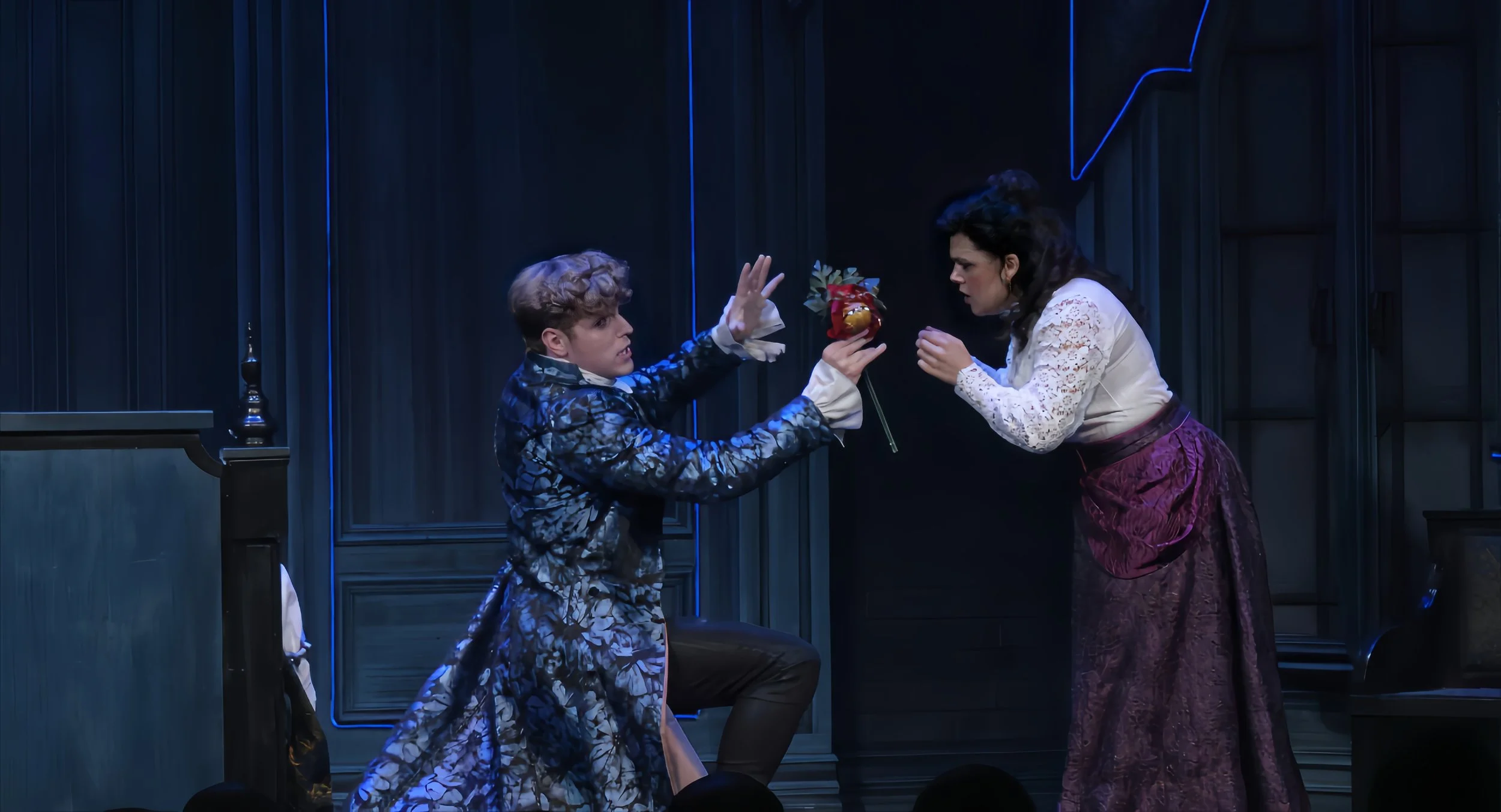 Two actors on stage, one kneeling and holding a red flower, the other reaching out towards it, in a theatrical performance with dark background.