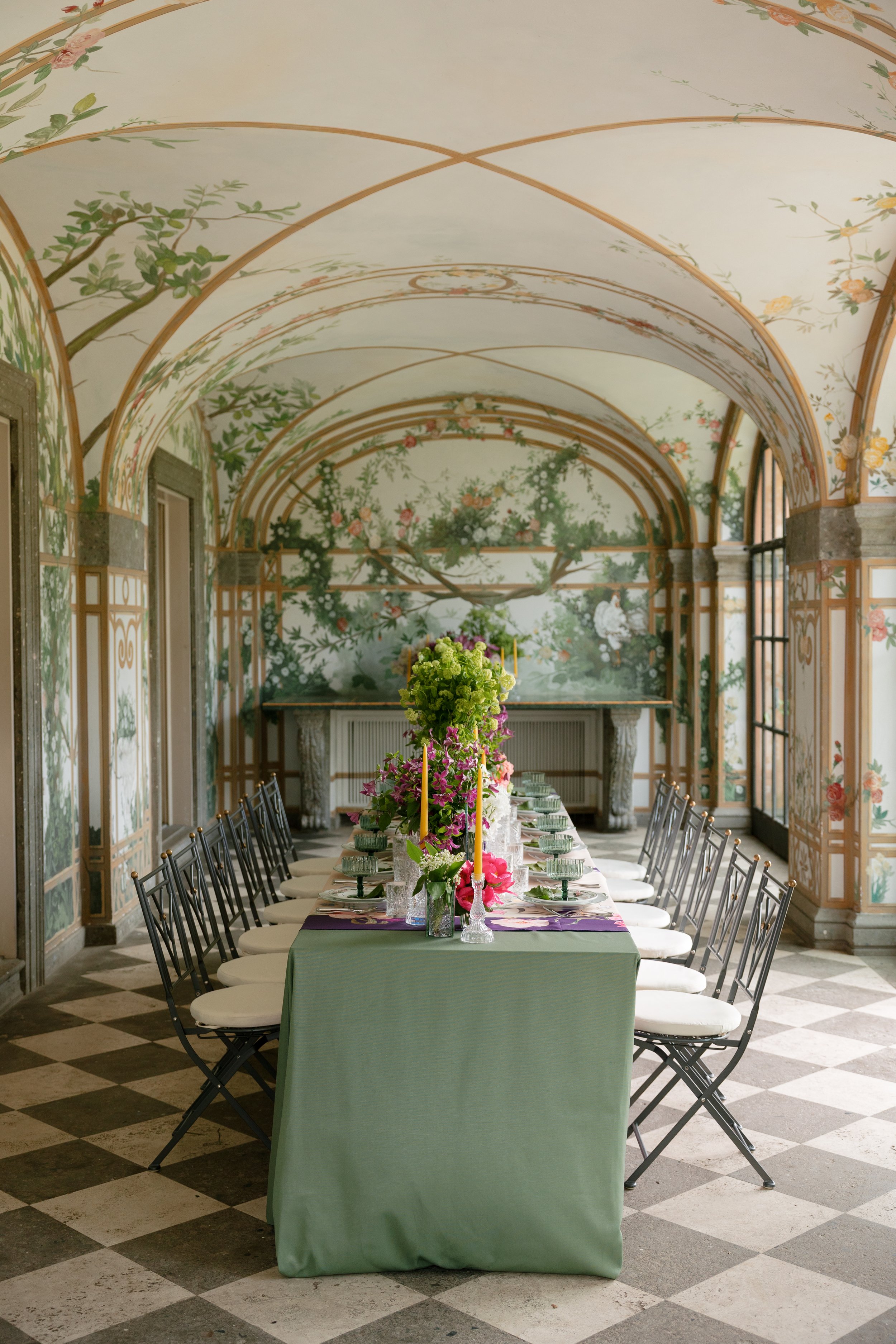 Elegant indoor dining room with a long table decorated with flowers, surrounded by black chairs with white cushions. The room features a painted, floral, arched ceiling and large windows, with a checkered black and white floor.